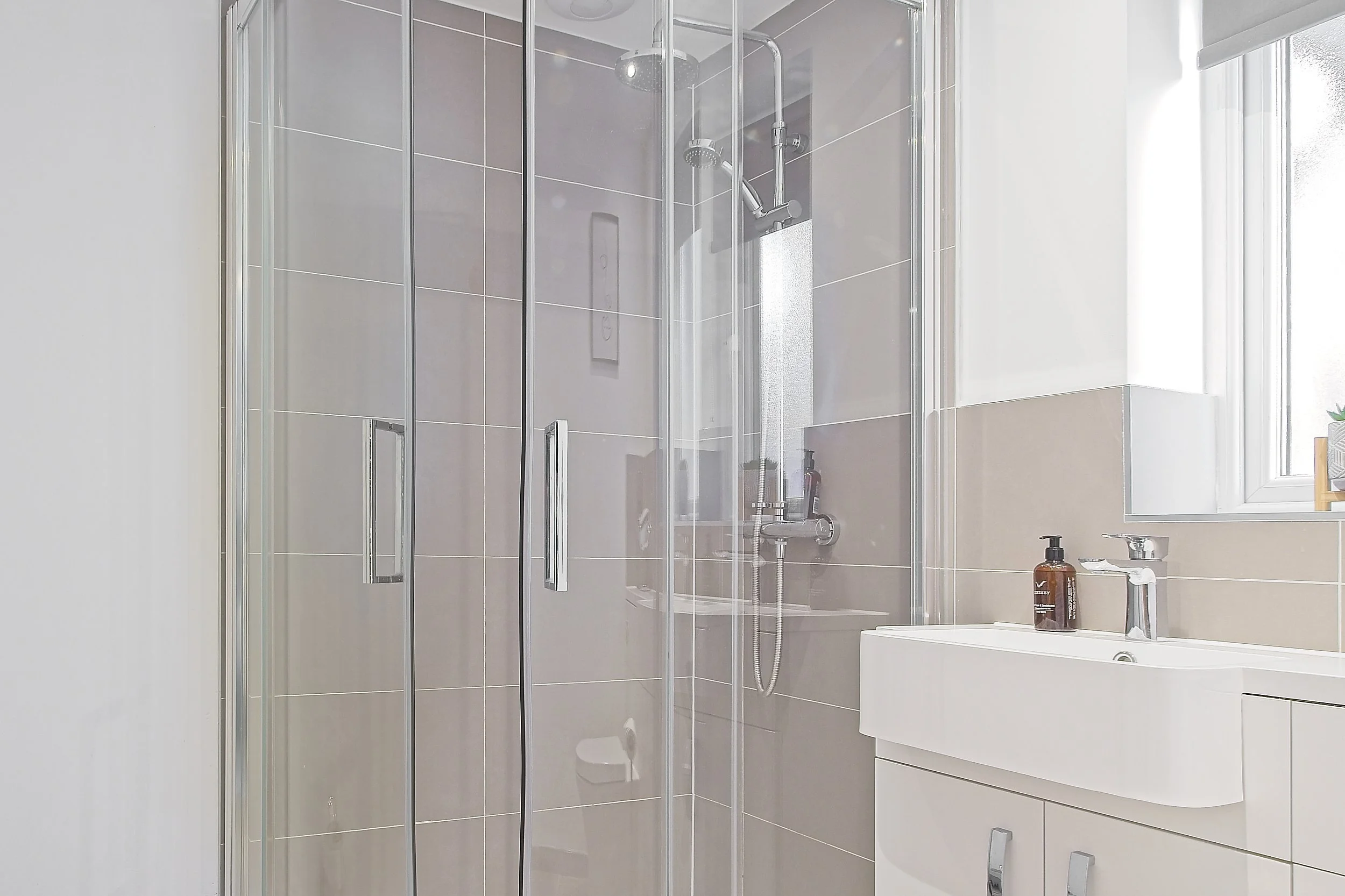 A modern bathroom shower with glass doors, a showerhead, and beige tiles, adjacent to a white vanity with a brown soap dispenser and a bottle.