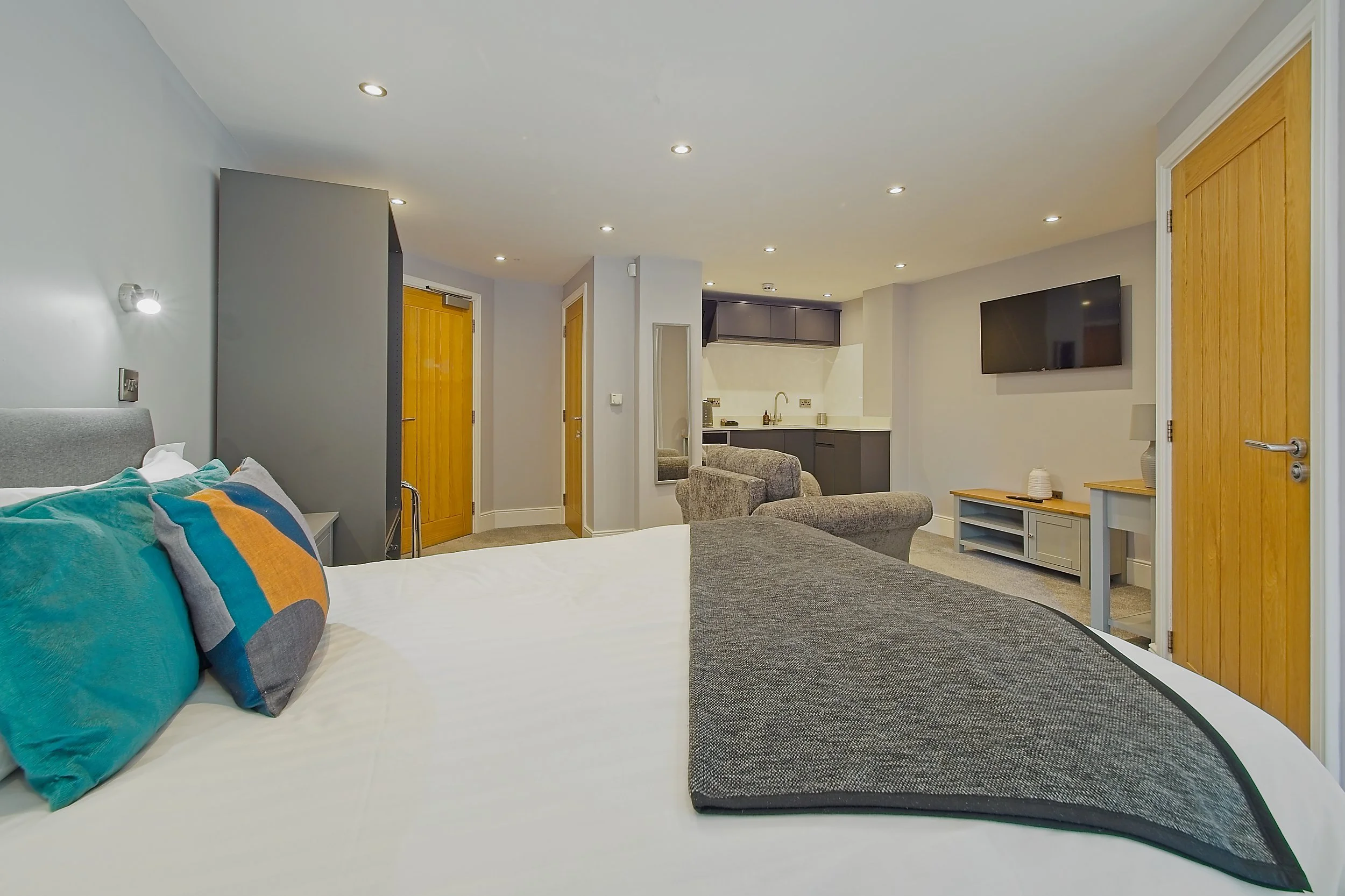 A modern living room with a bed, colorful pillows, two armchairs, a wall-mounted TV, and a small kitchenette area with gray cabinets.
