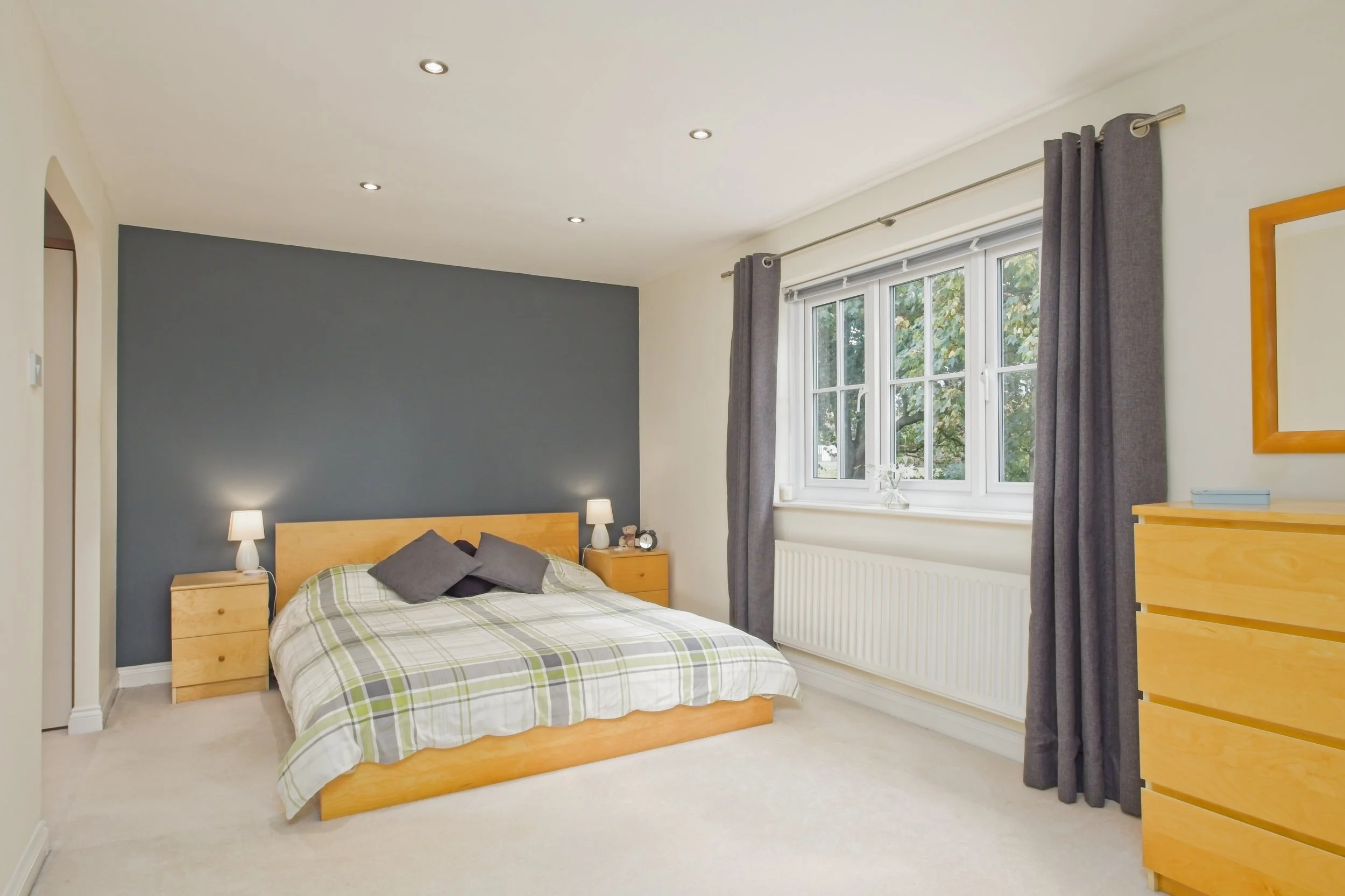 A bedroom with a wooden bed frame, a plaid bedspread, two dark gray pillows, two wooden nightstands with lamps, a window with gray curtains, and a yellow dresser, all under a white ceiling with recessed lighting.
