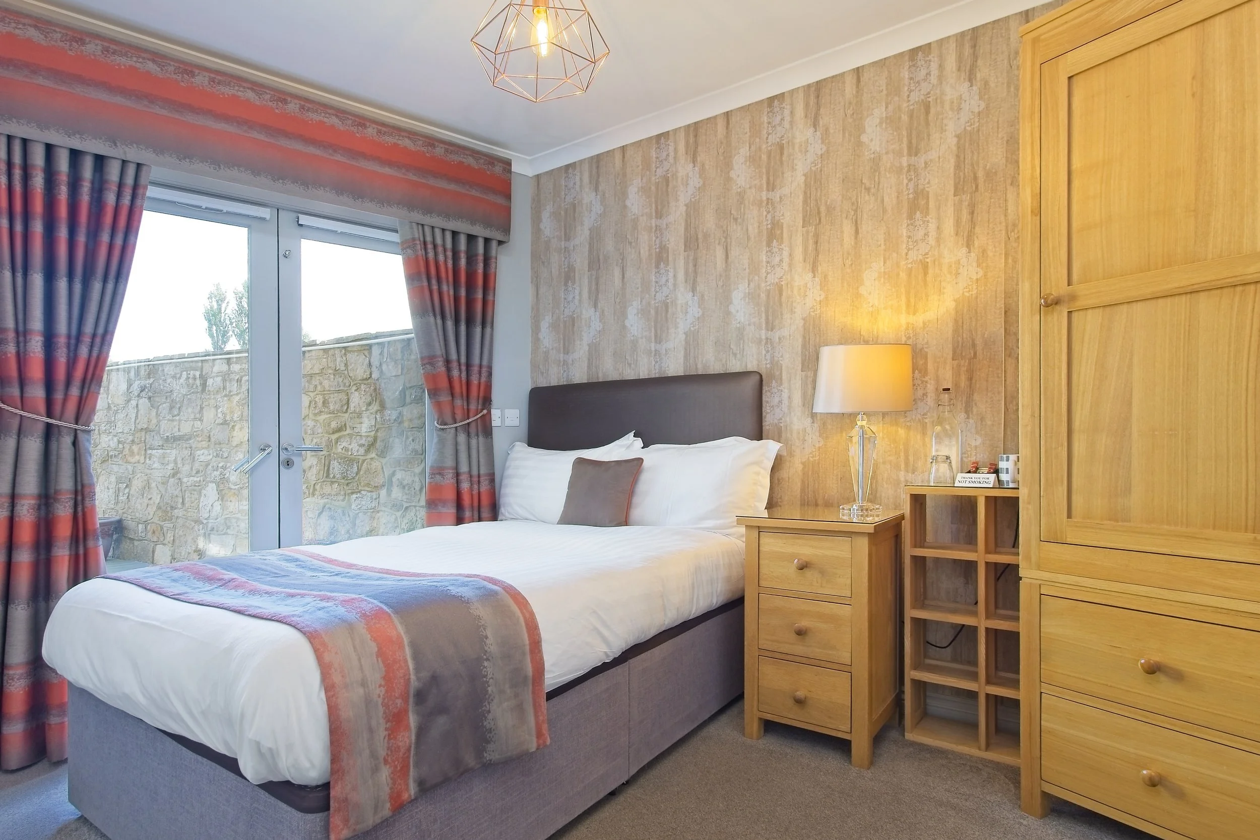 A bedroom with a bed, bedside table, lamp, wooden cabinet, and window with curtains.