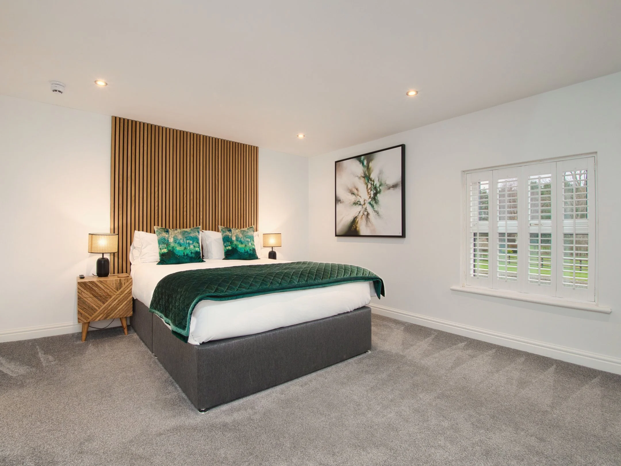 A modern bedroom with a bed that has white linens, teal and blue decorative pillows, a dark green quilt, and a gray upholstered base. On either side of the bed are matching wooden nightstands with black lamps. The headboard is a vertical slat wooden 