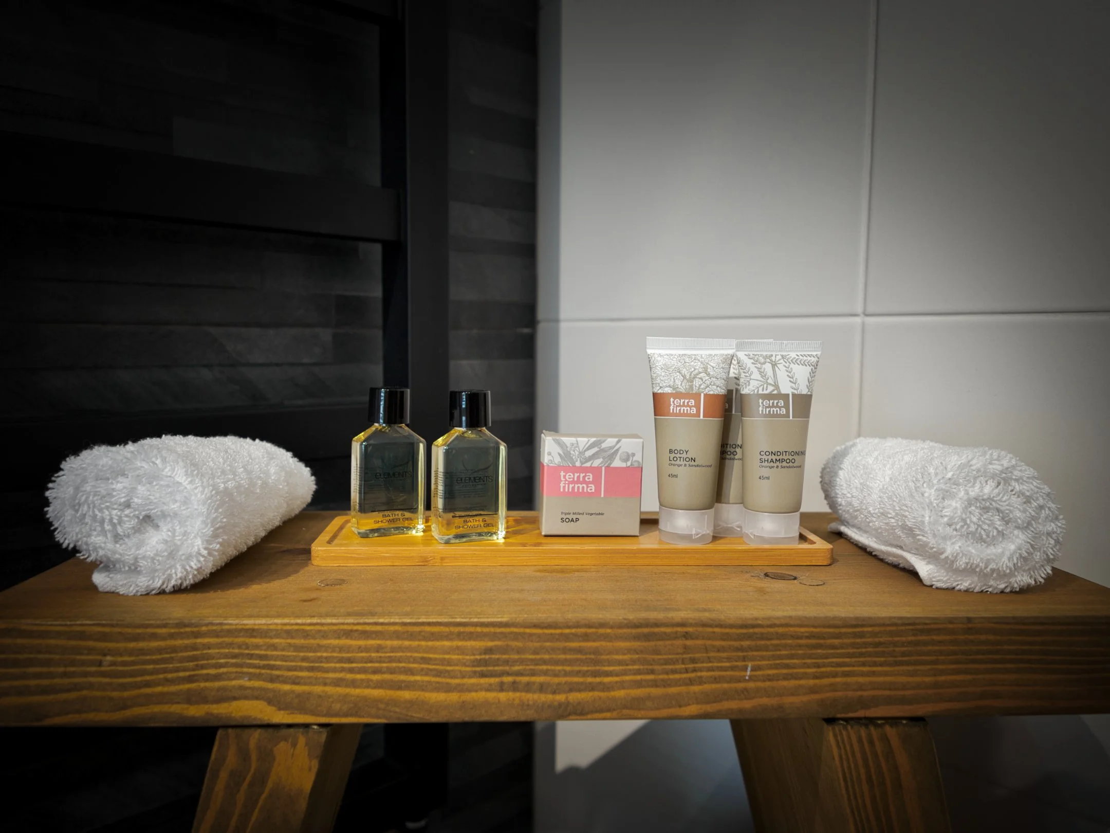 Bathroom counter with toiletries including shower gel, body lotion, soap, shampoo, and two rolled white towels on a wooden surface.