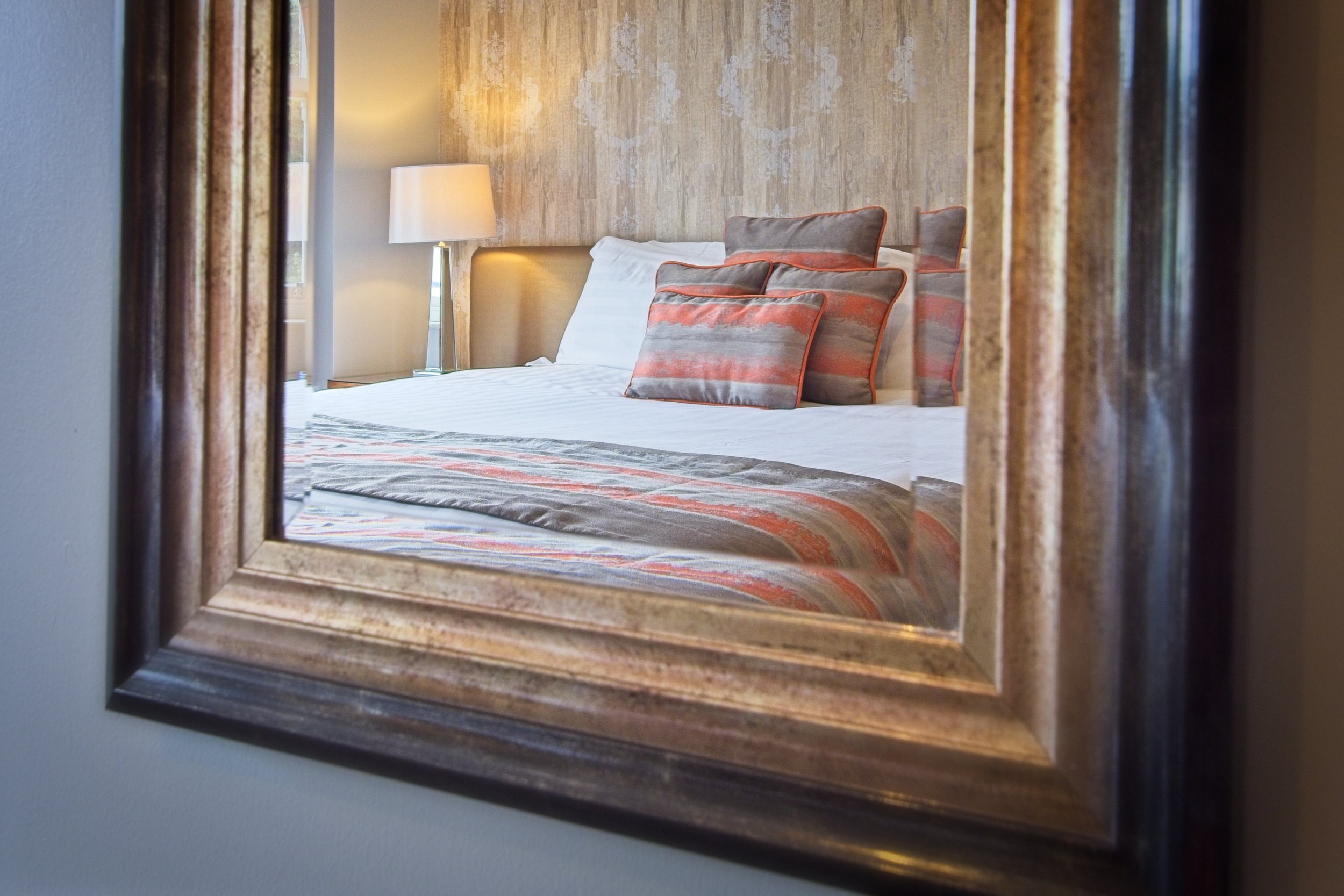 Reflection of a bedroom in a mirror showing a bed with pillows and a lamp on a bedside table.
