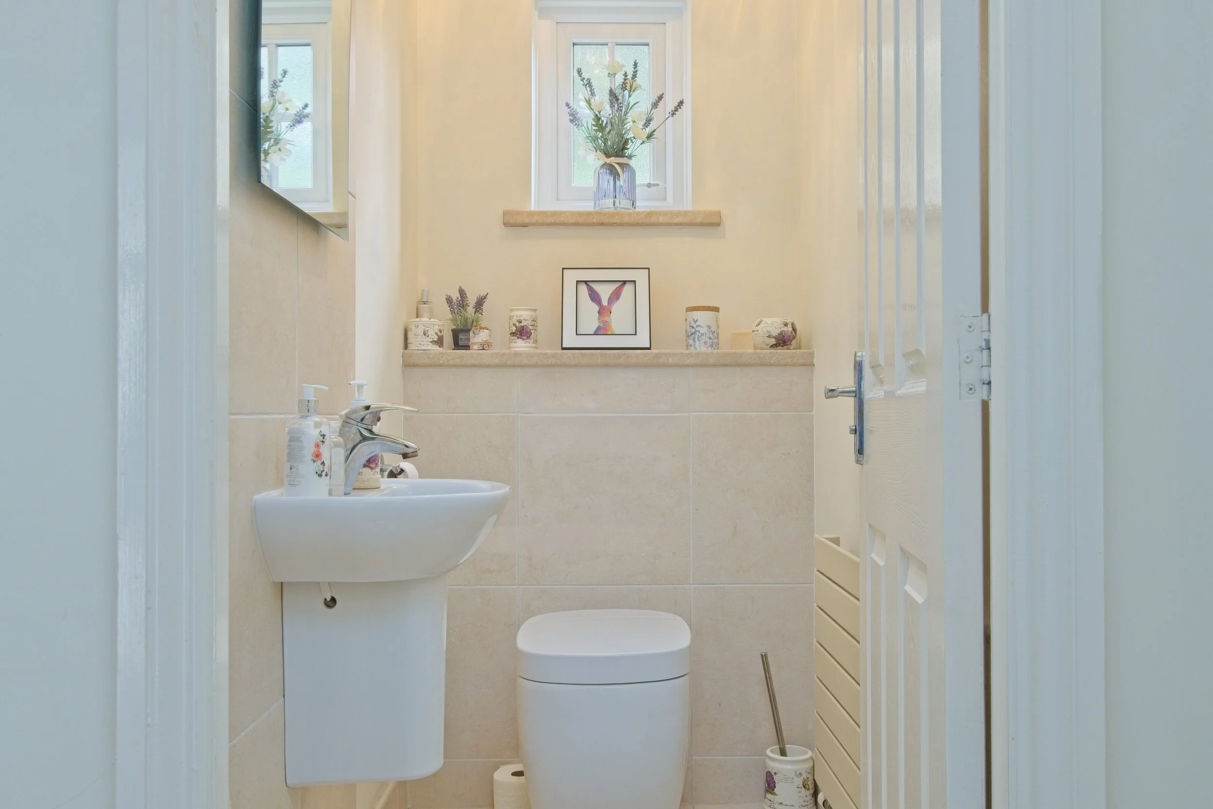 A small bathroom with a sink, toilet, and decorated shelf with plants and artwork, illuminated by natural light from a window.