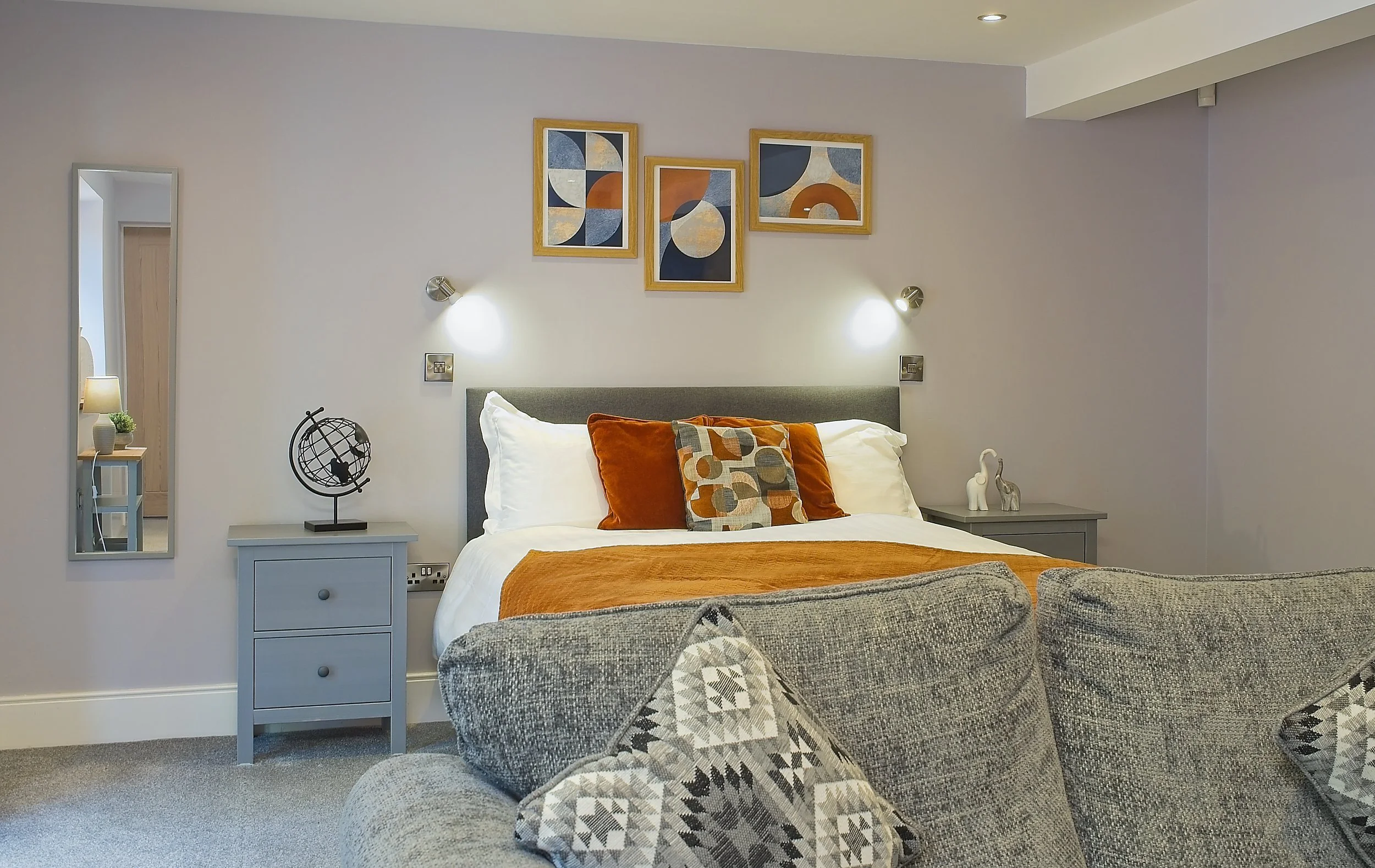 A bedroom with a bed, decorative pillows, and wall art. The wall art includes four framed abstract paintings. There are bedside tables on either side, with lamps and decorative objects. A mirror is visible on the left side. The room has neutral tones with accents of orange and gray.