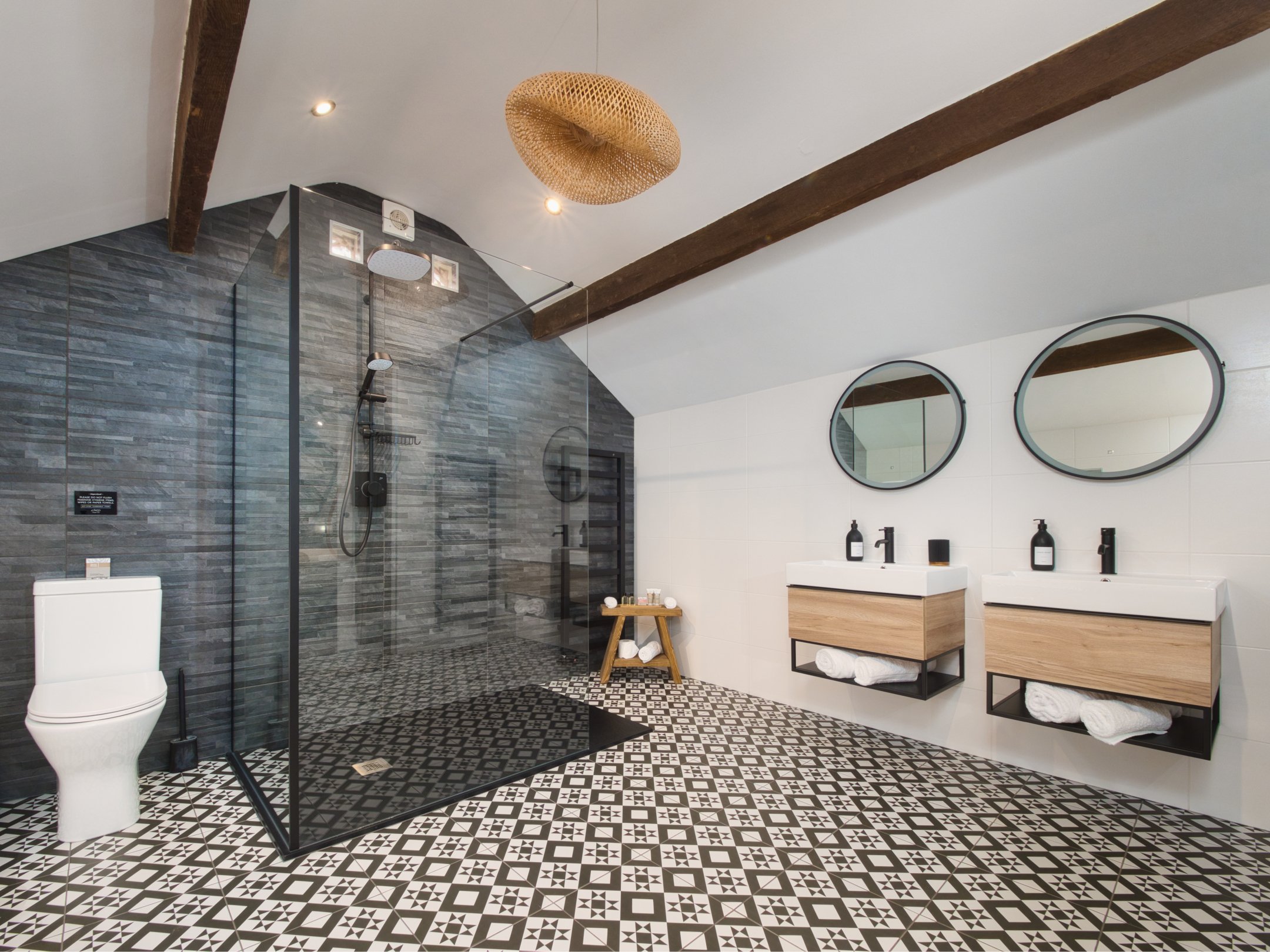 Modern bathroom with black and white patterned floor, a walk-in shower with gray stone tiles, a white toilet, and a double vanity with black fixtures and rounded mirrors.
