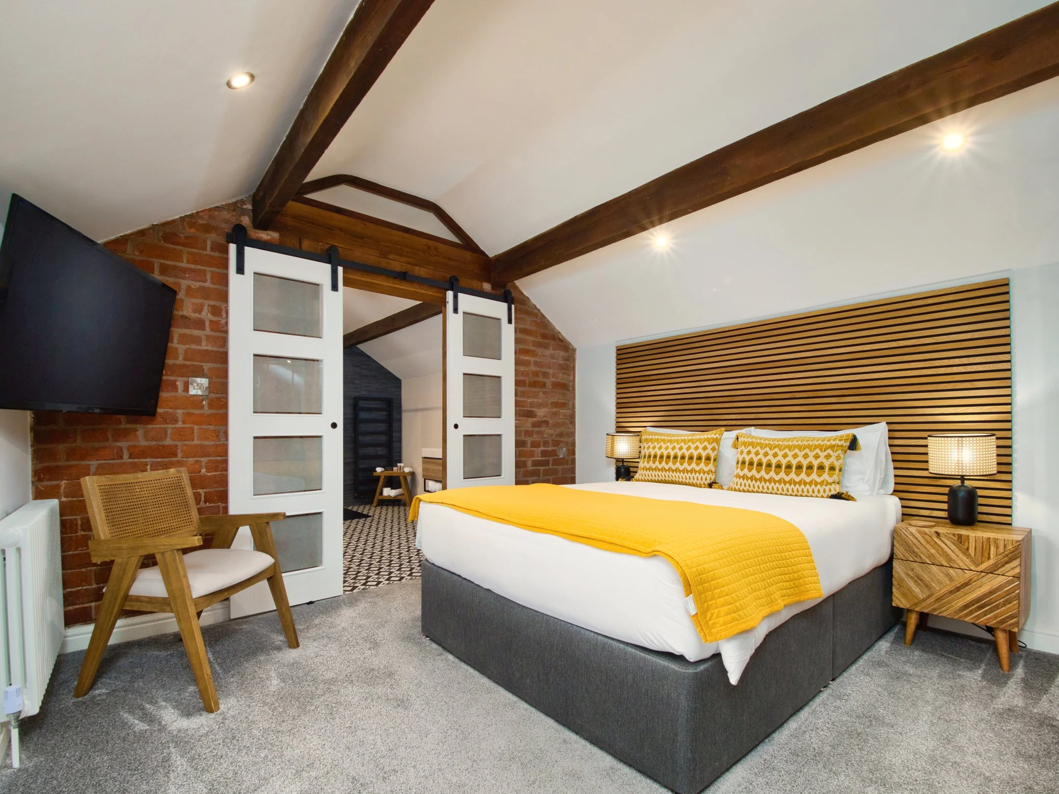 A bedroom with a gray bed frame, white bed linens, and yellow blankets and pillows. Wooden accents, including a headboard made of horizontal slats and a side table. A pair of bedside lamps with black bases and woven shades. A white sliding barn door 