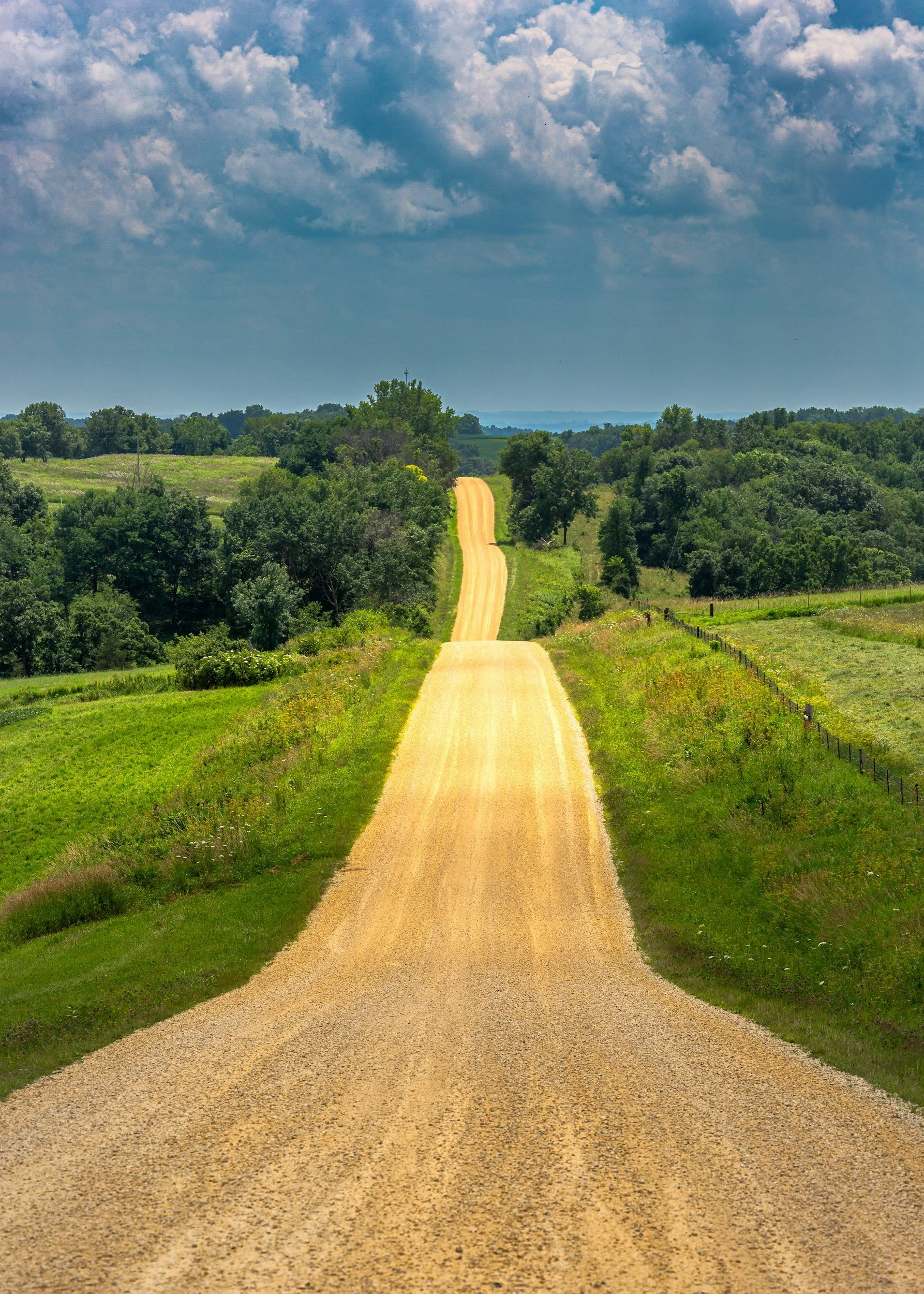 A dirt road running through green fields and trees under a partly cloudy sky.