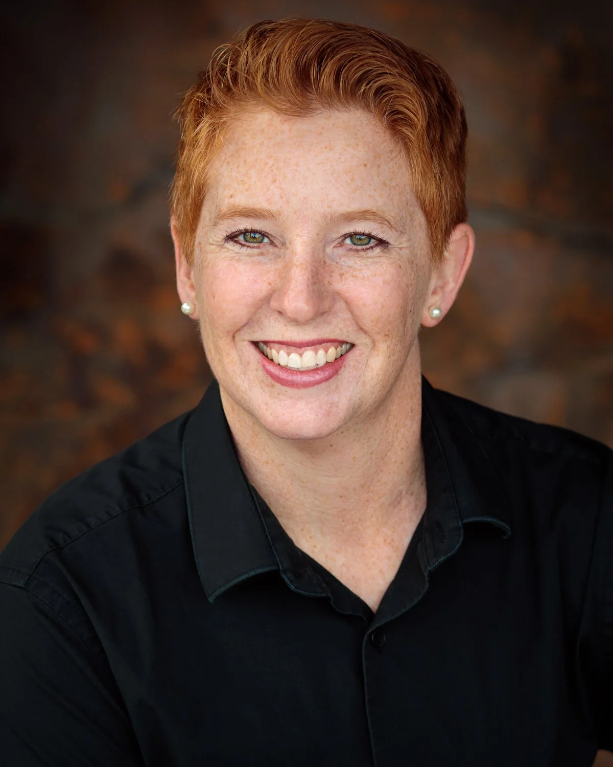Close-up portrait of Julie Lucas smiling with short red hair, light skin with freckles, pearl earrings and a black shirt.