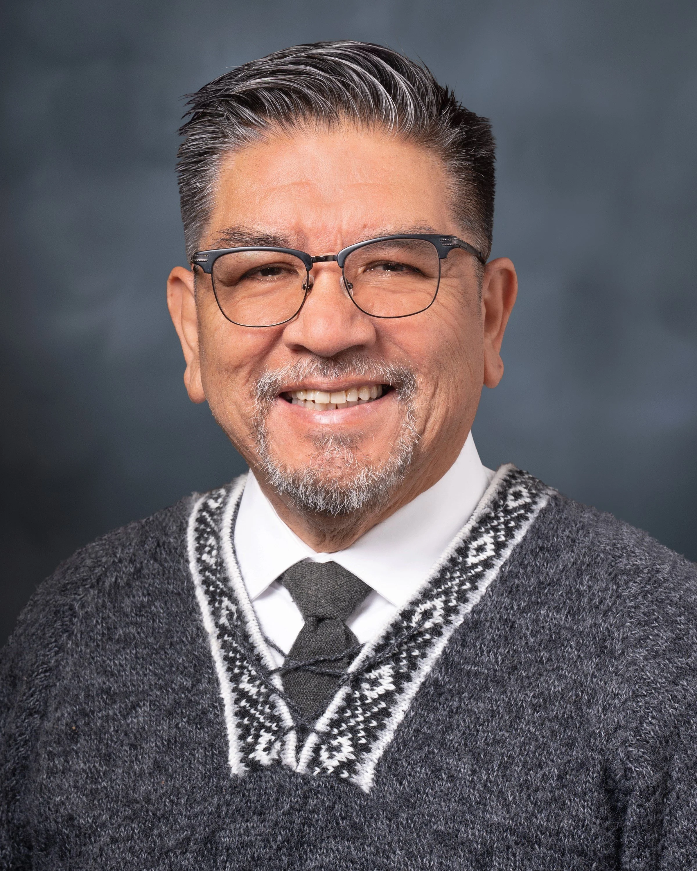 Close-up of Pablo Obregon, smiling with glasses, salt-and-pepper hair, and a beard, wearing a white shirt, dark tie, and patterned gray cardigan.