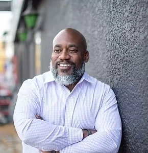 Portrait of Justin Terrell leaning against a wall in a checkered shirt with arms crossed and a warm smile.