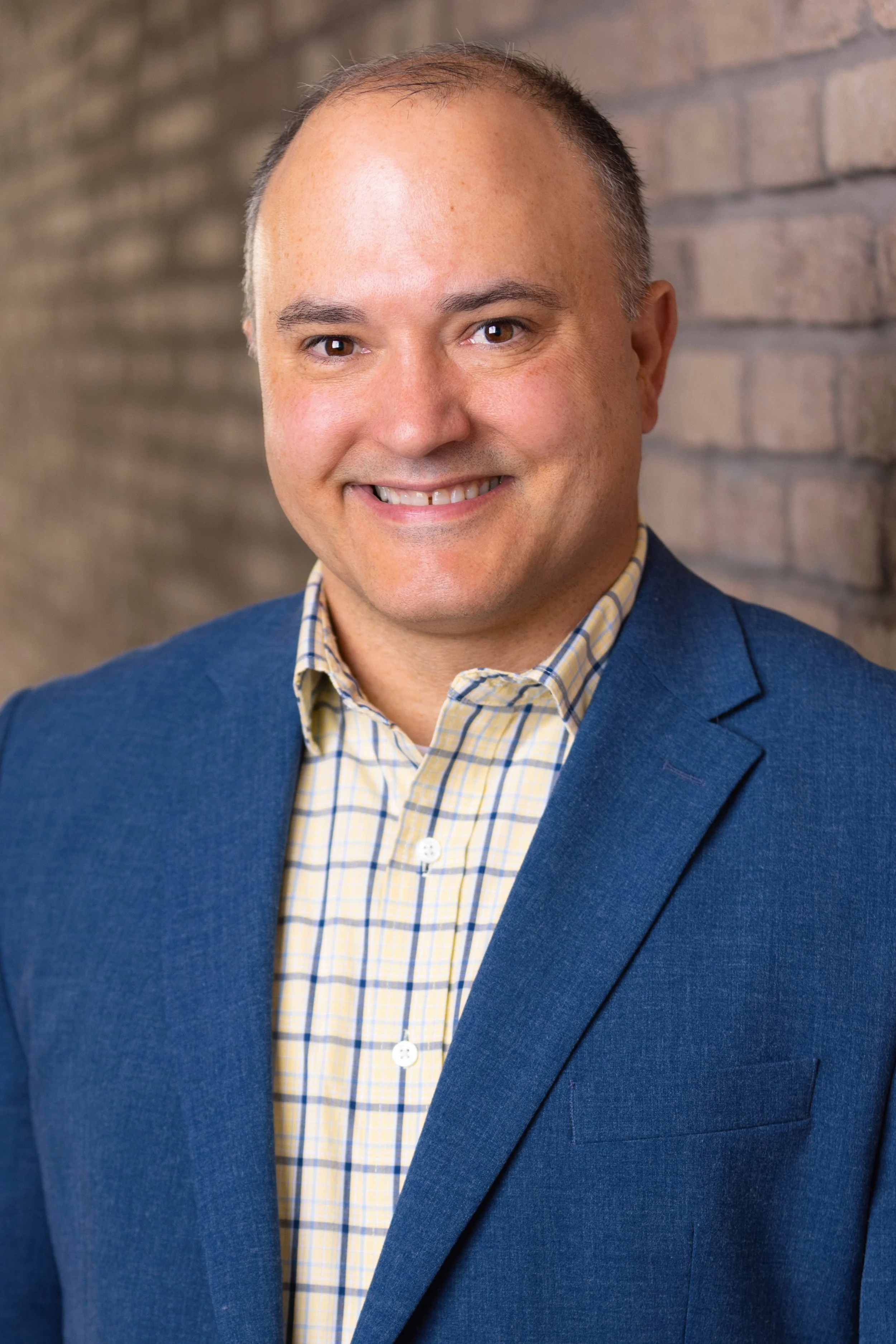 Portrait of Rick Bauerly in front of a brick wall wearing a blue suit and checkered shirt. 