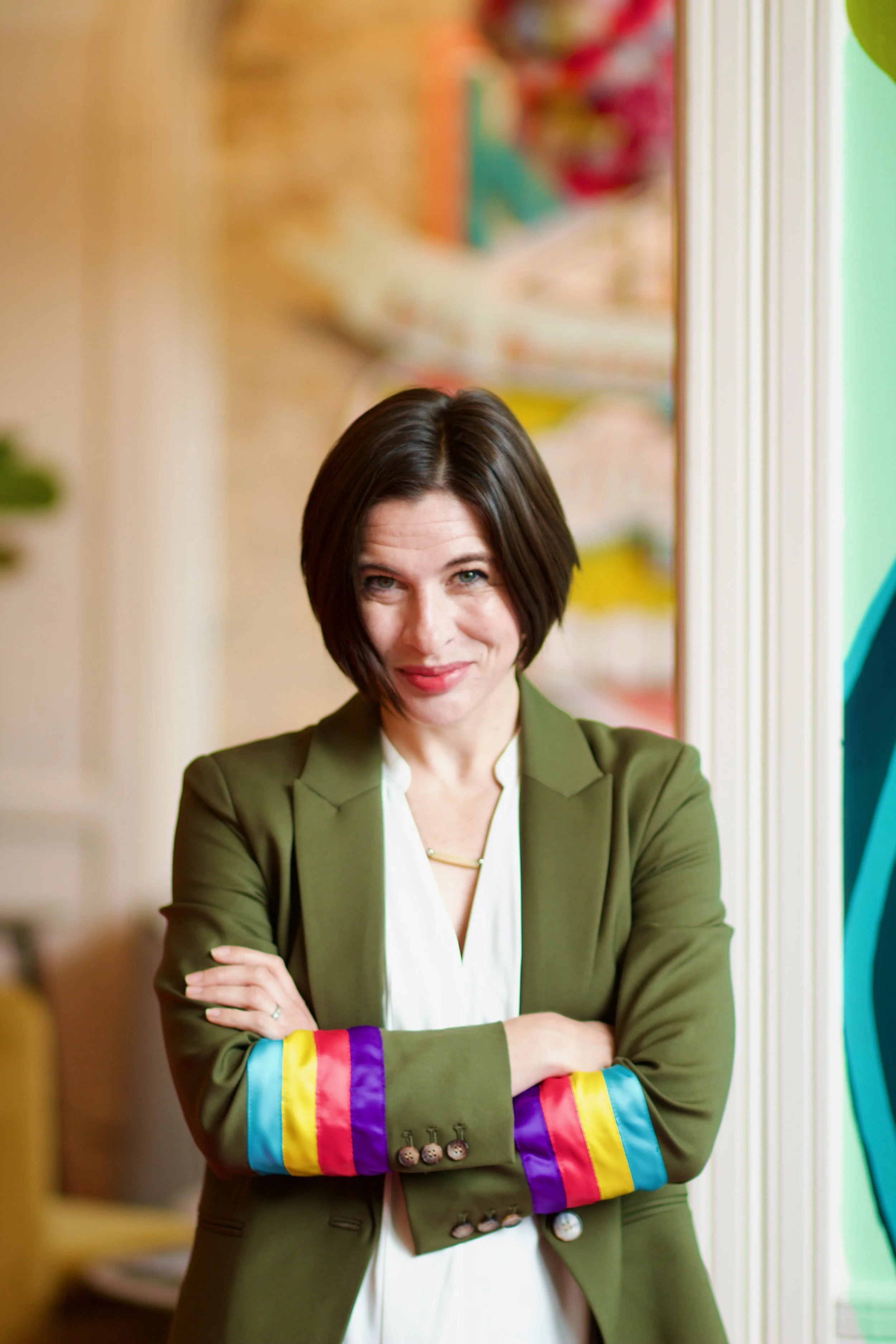 Portrait of Jenna Ray standing in a hallway smiling in an olive green blazer cuffed with brightly colored stripes.