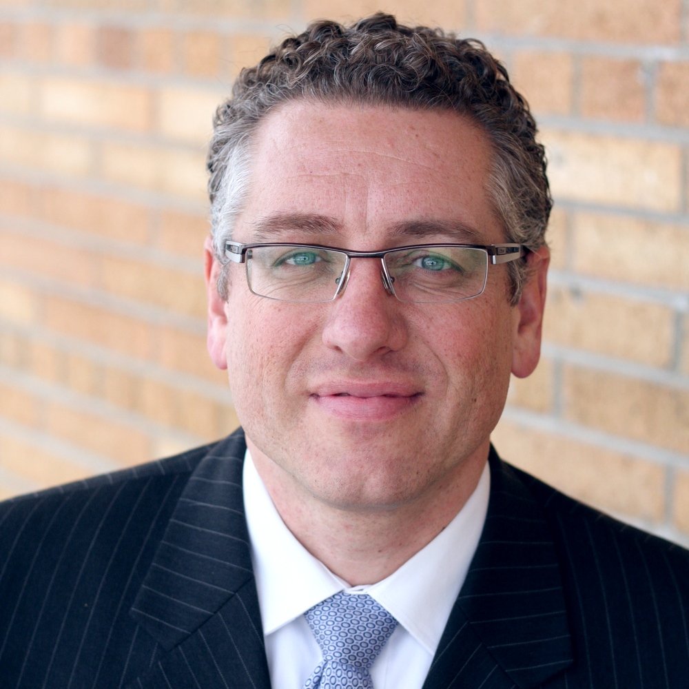A man with curly hair and glasses wearing a pinstripe suit, white shirt, and light patterned tie, standing in front of a brick wall.