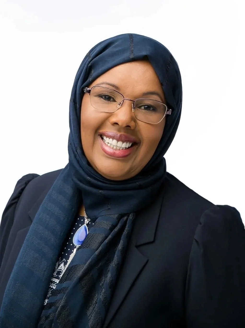 Portrait of Fatoun Ali with a radiant smile, wearing rectangular wire-rimmed glasses and a navy blue head scarf.