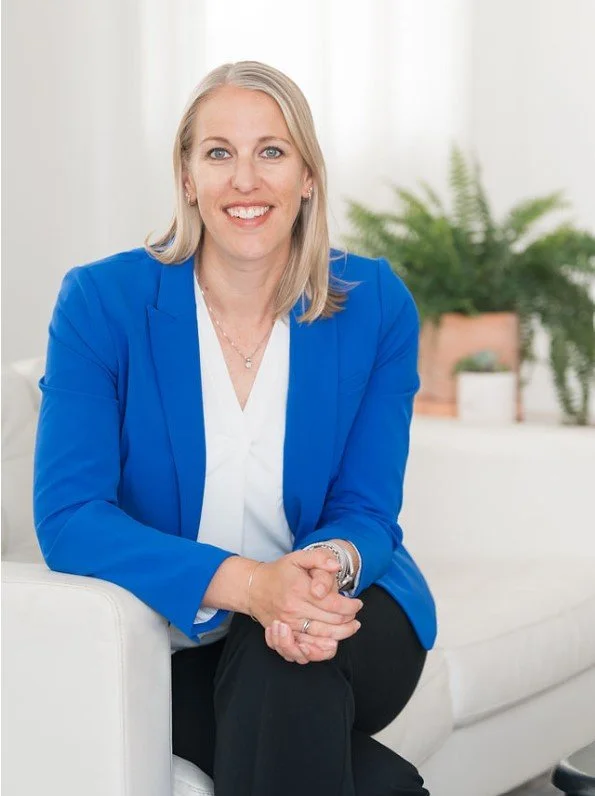 A smiling woman with blonde hair wearing a bright blue blazer, white blouse, and black pants, sitting on a white couch in a bright, modern room with a potted plant in the background.