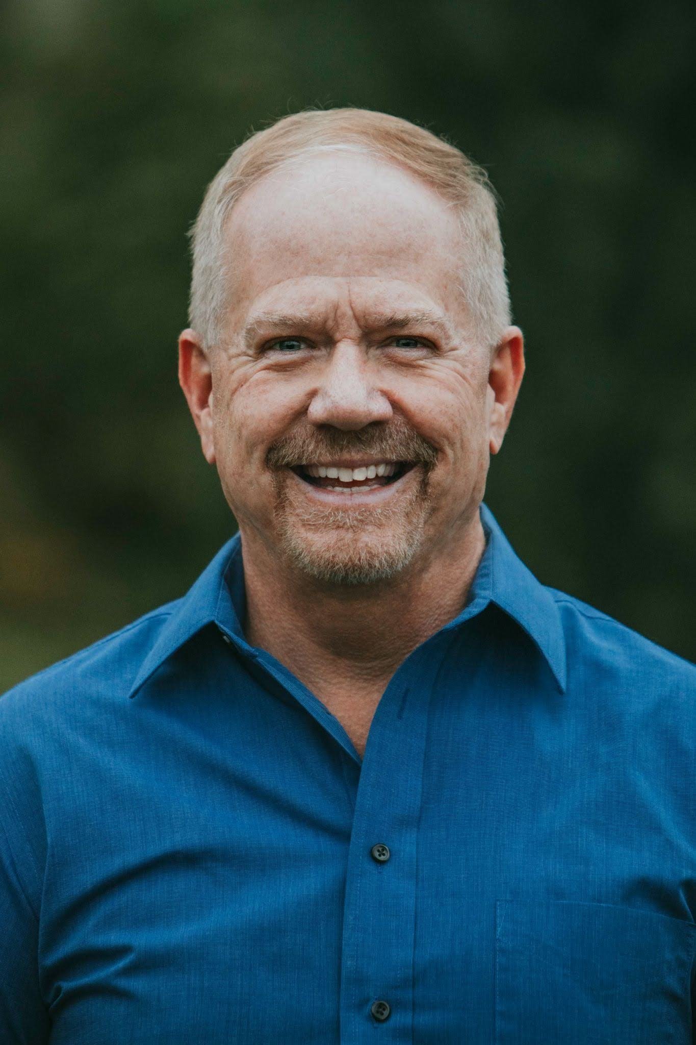 Steve Eng, smiling with short-cropped hair, blue eyes, and a goatee, wearing a blue button-up shirt, standing outdoors with a blurred green background.