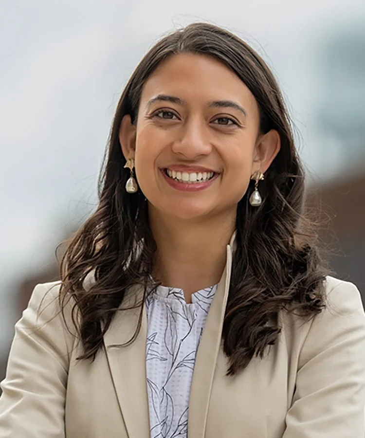 Portrait of Benya Kraus, smiling broadly with curly brown hair, large teardrop pearl earrlings, and a tan blazer.
