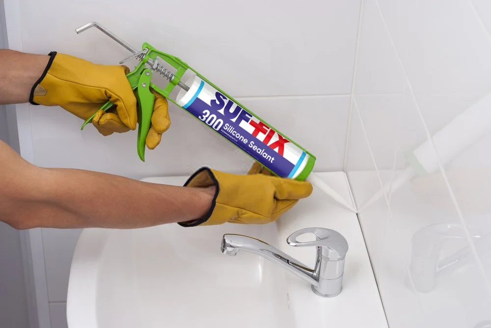 Hands wearing yellow gloves holding a silicone sealant caulking gun above a bathroom sink.