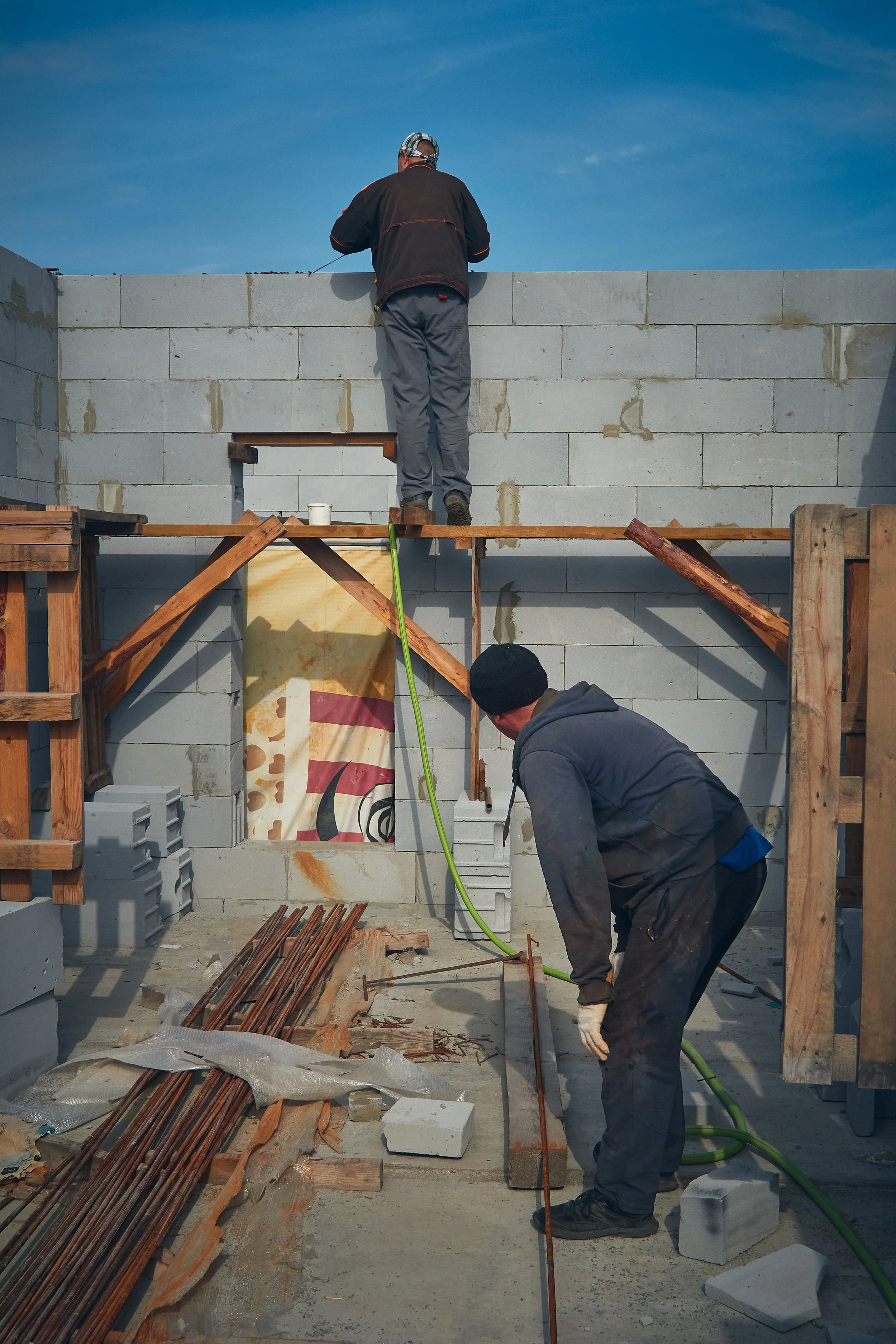 Two construction workers are building a cinder block wall outdoors. One worker is standing on a wooden platform laying blocks, while another is kneeling on the ground, working on the foundation. Building supplies and tools are around them, with a clear blue sky above.