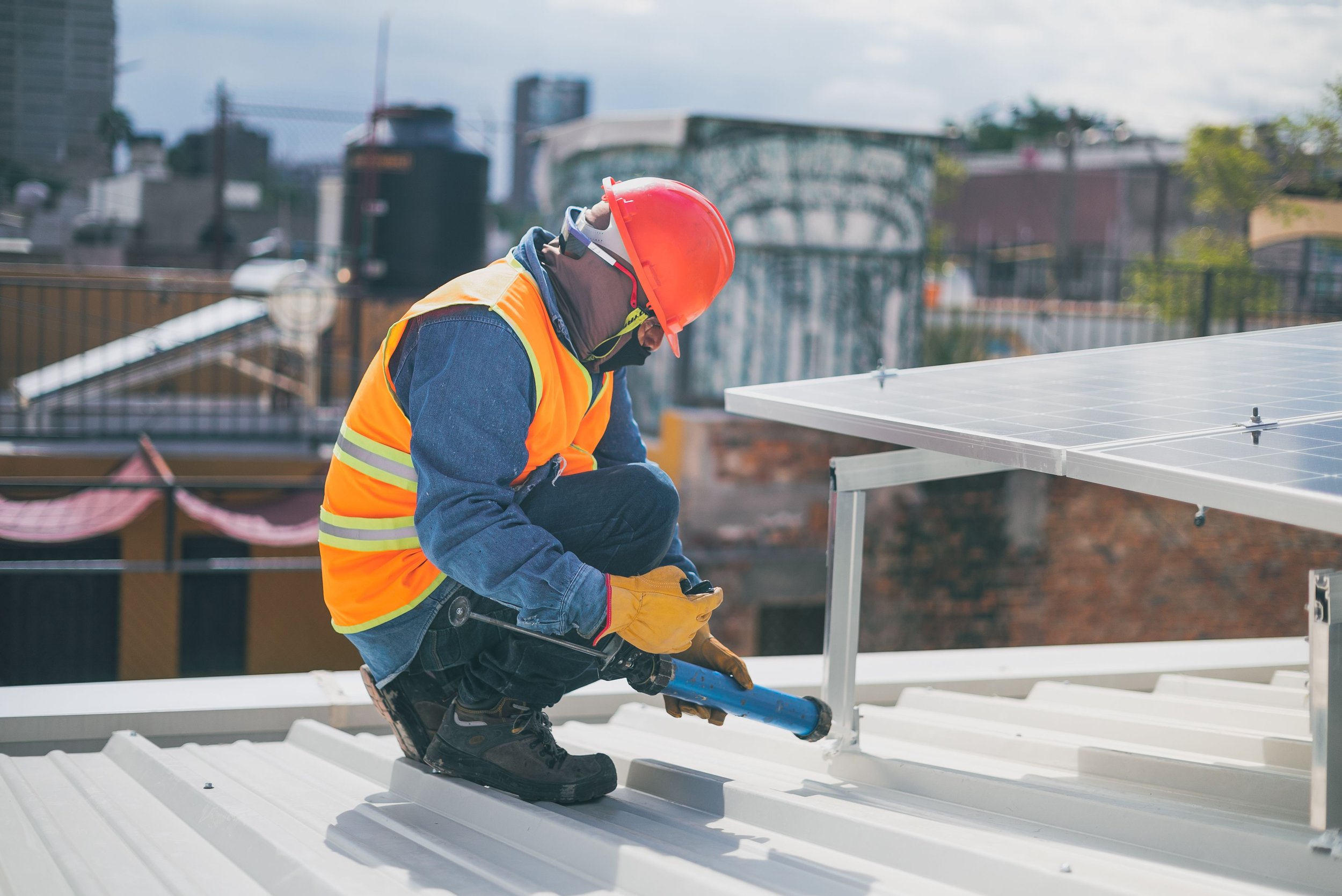 Construction worker installing solar panels on a roof, wearing safety gear including a red helmet, yellow safety vest, gloves, and safety glasses, with an urban cityscape background.