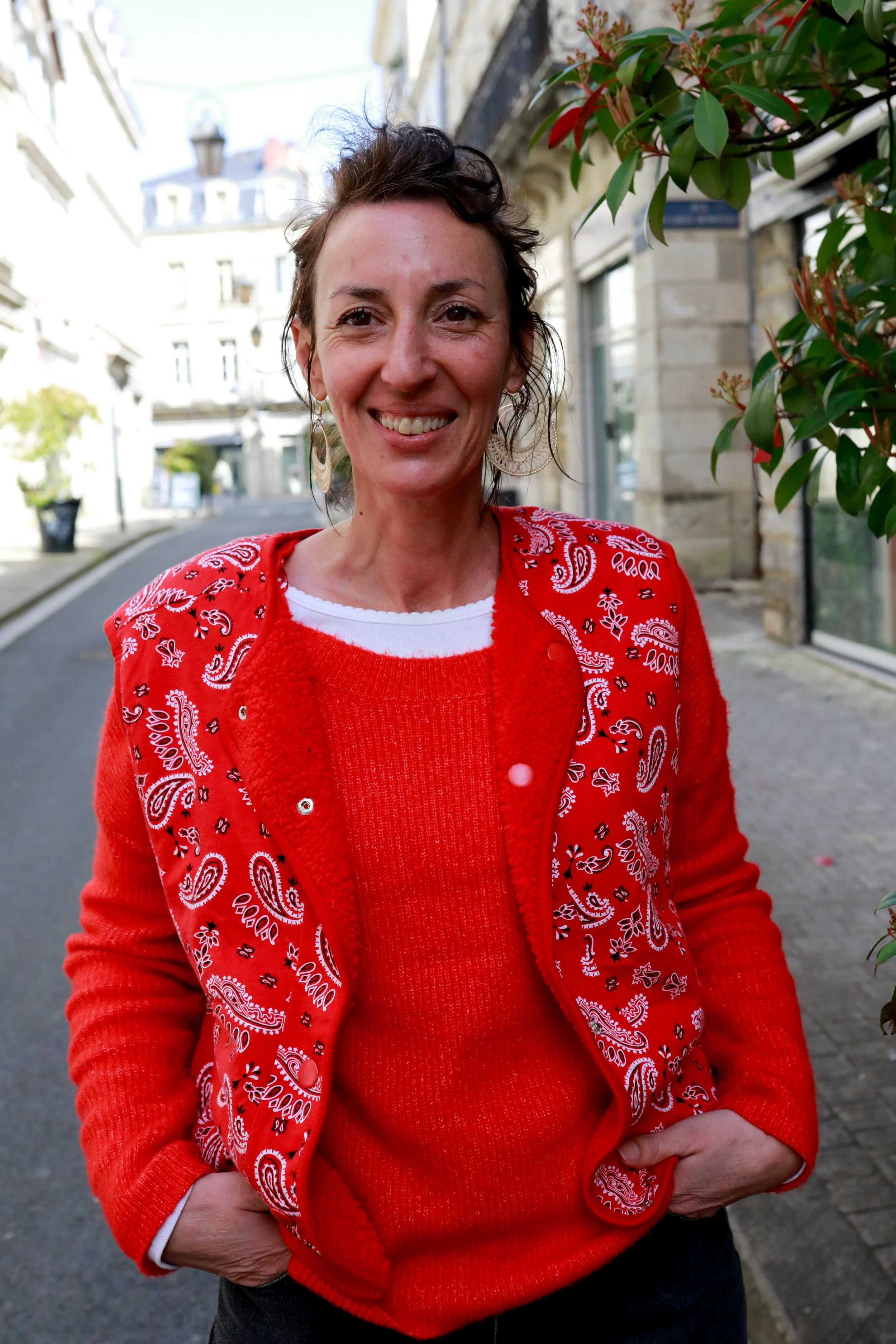 Femme souriante portant un pull rouge et une veste rouge à motifs bandana, dehors dans une rue urbaine avec des bâtiments en arrière-plan.