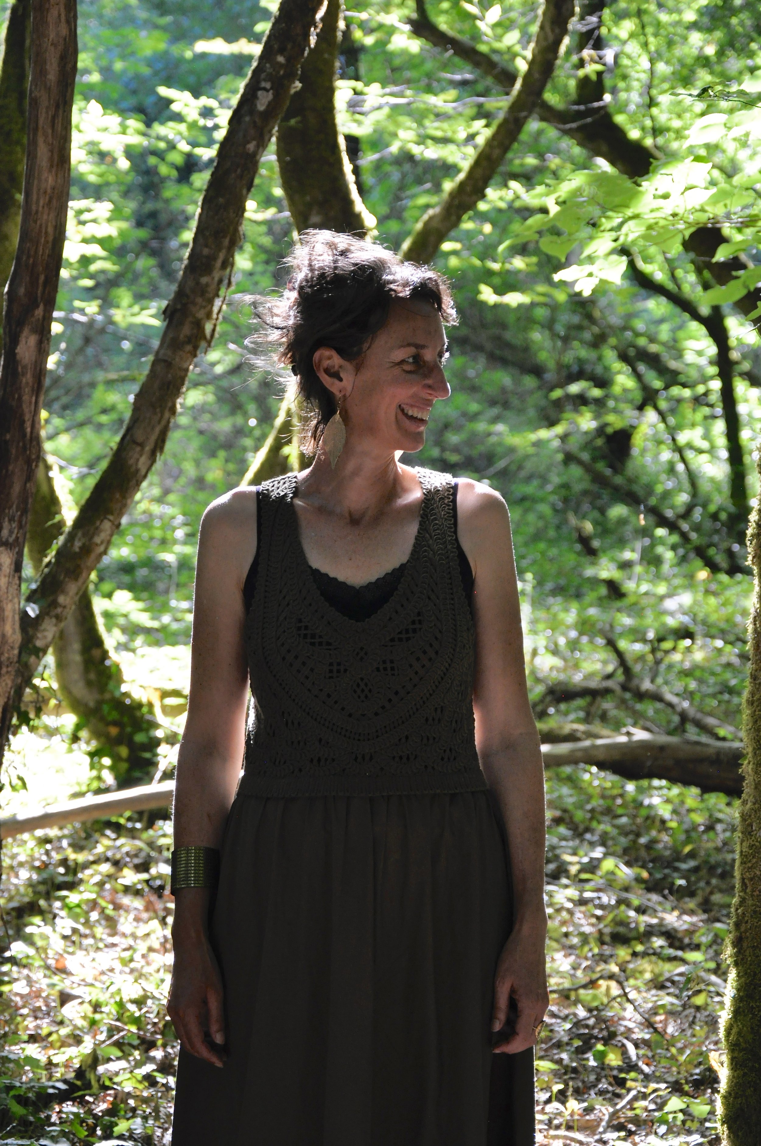 Une femme souriante dans une forêt verdoyante, portant une robe noire sans manches et une boucle d'oreille unique.