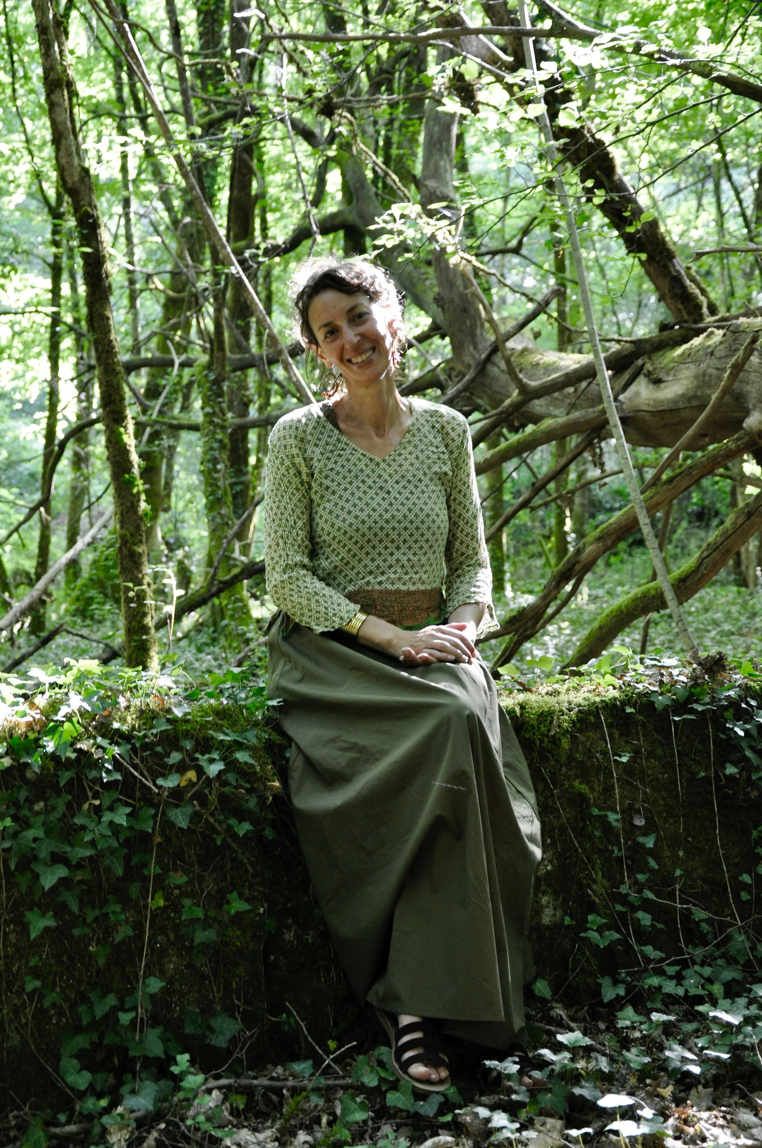 Femme assise sur un tronc d'arbre dans une forêt verdoyante, portant une robe longue et des sandales à la mode, souriant à la caméra.