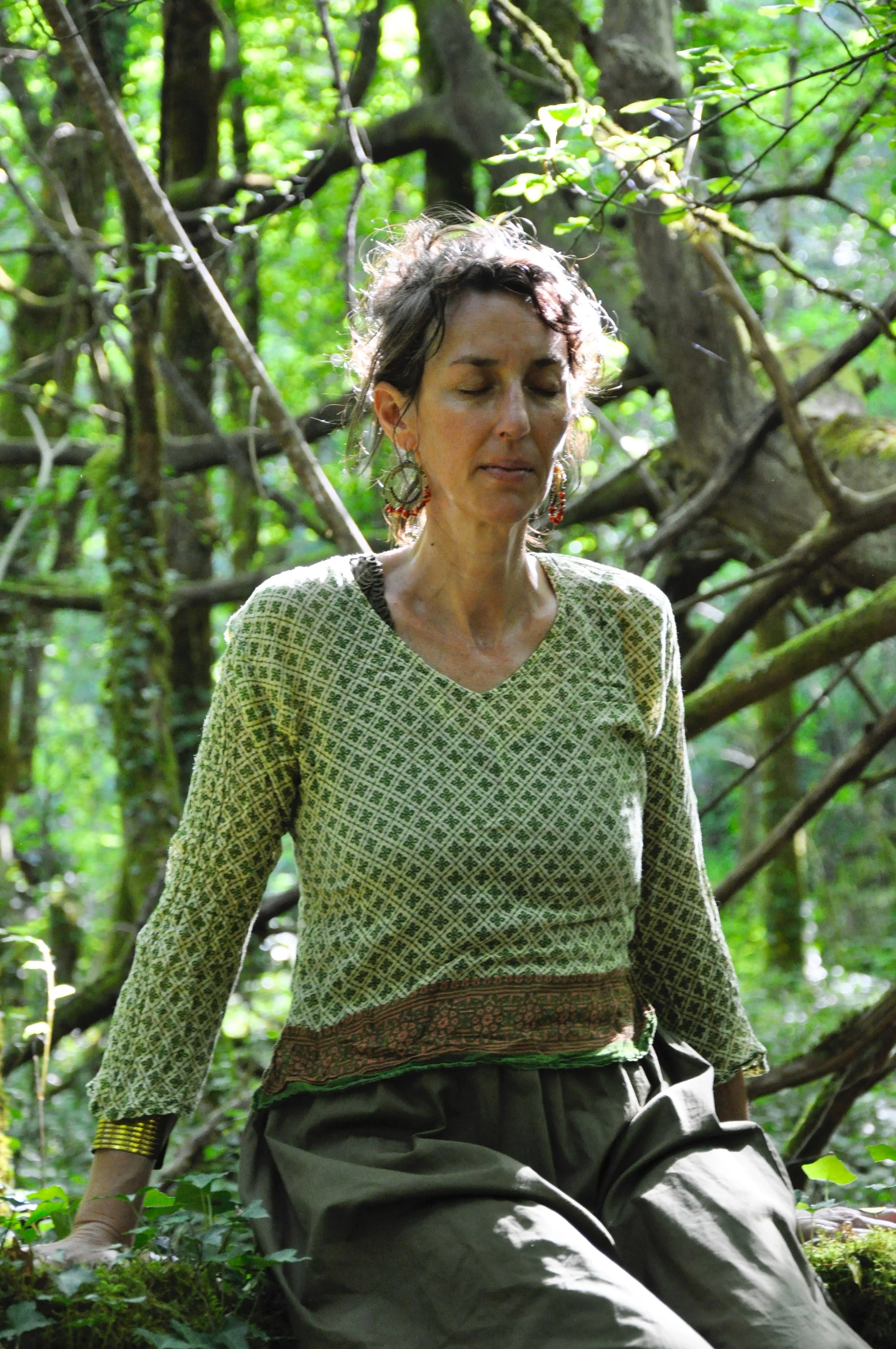 Une femme assise dans la forêt, yeux fermés, portant un vêtement à motif vert et une jupe en tissu avec des détails traditionnels. Environnement naturel verdoyant avec des branches d'arbres.