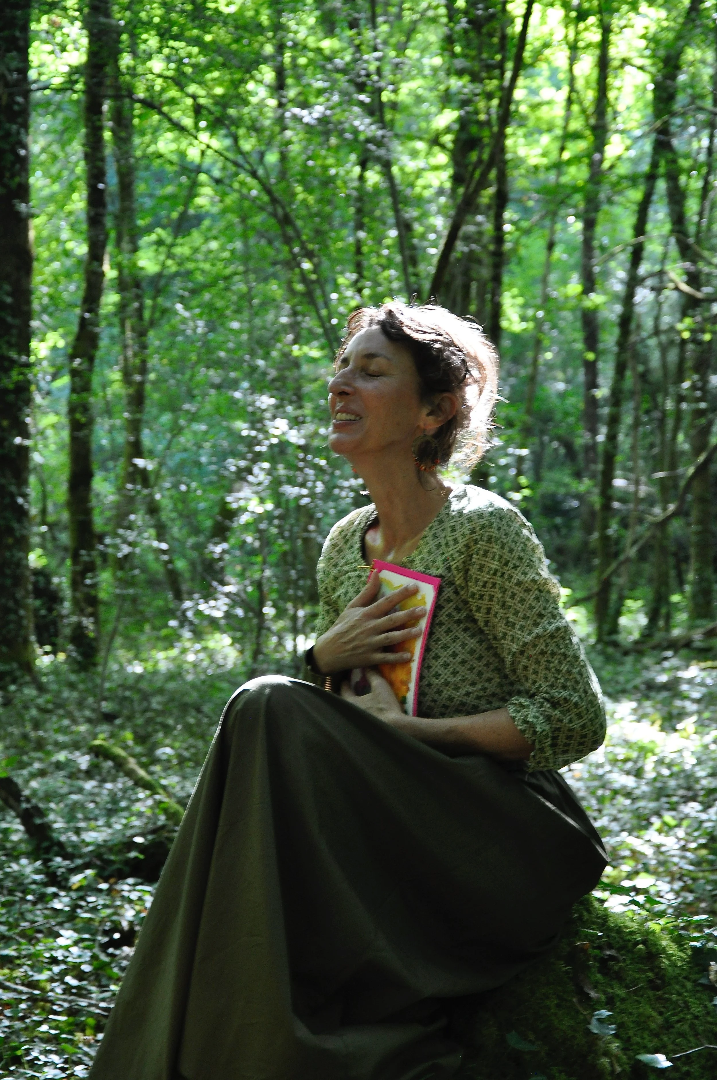 Une femme souriante, yeux fermés, dans une forêt dense et ensoleillée, tenant un livre contre sa poitrine.