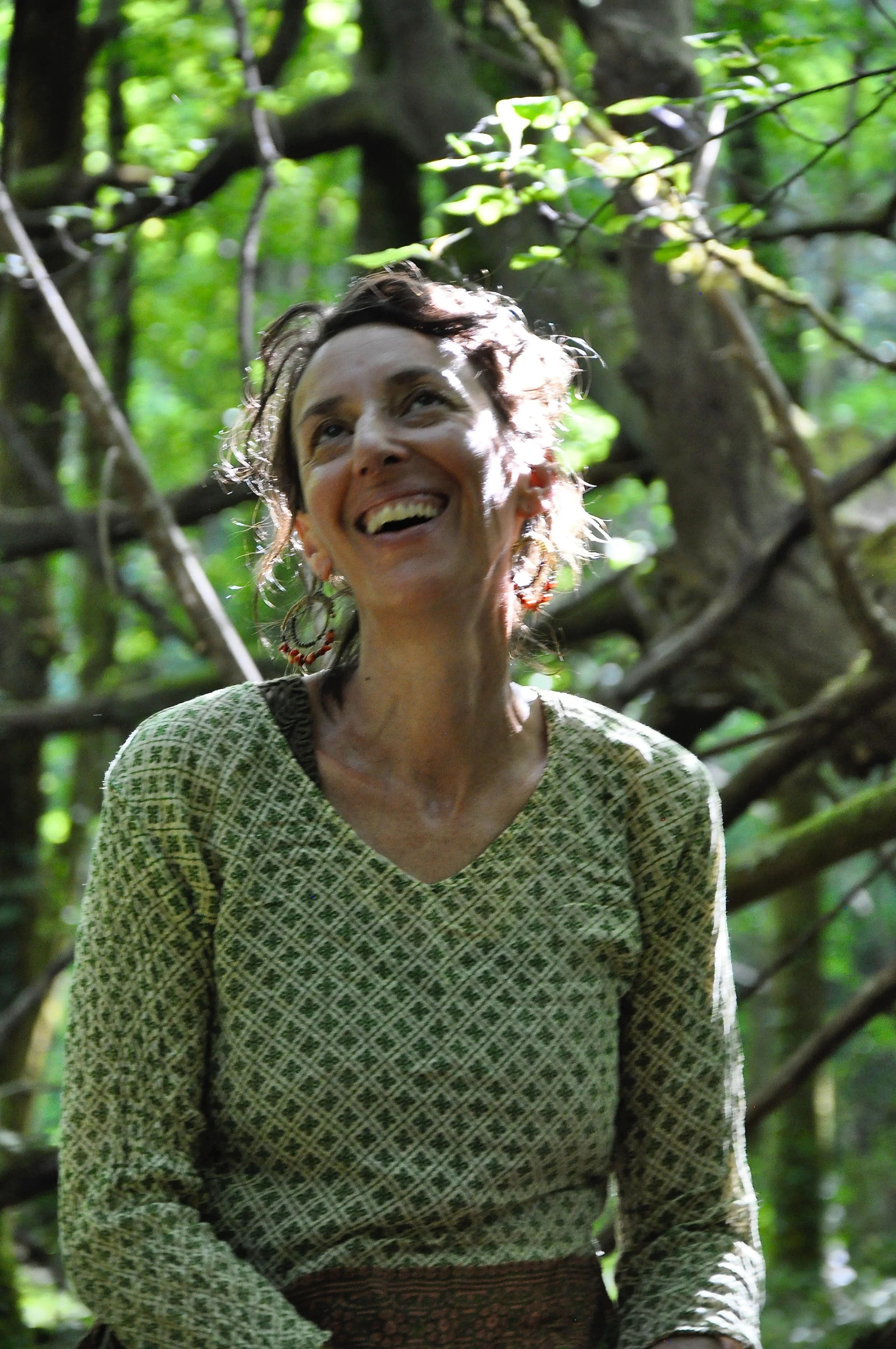 Femme souriante avec boucle d'oreille colorée, portant un vêtement en tissu vert à motif, dans une forêt avec beaucoup de feuillage vert