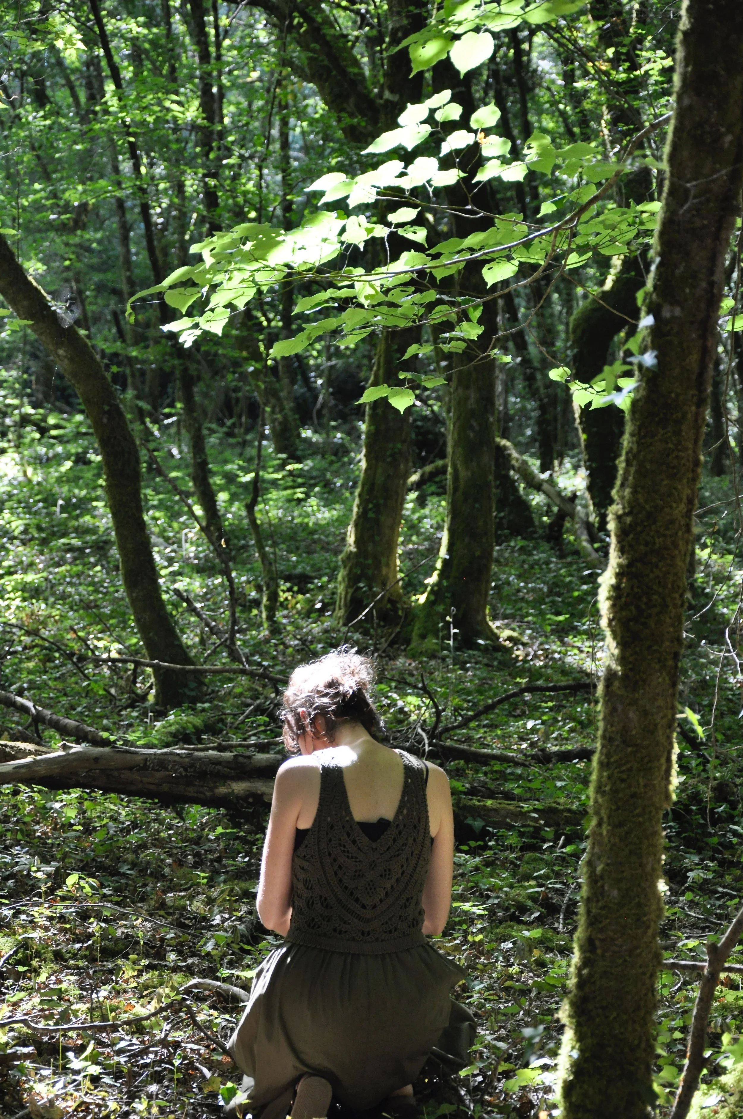 Une personne portant un débardeur noir assise ou accroupie dans une forêt dense, entourée d'arbres et de végétation verte.