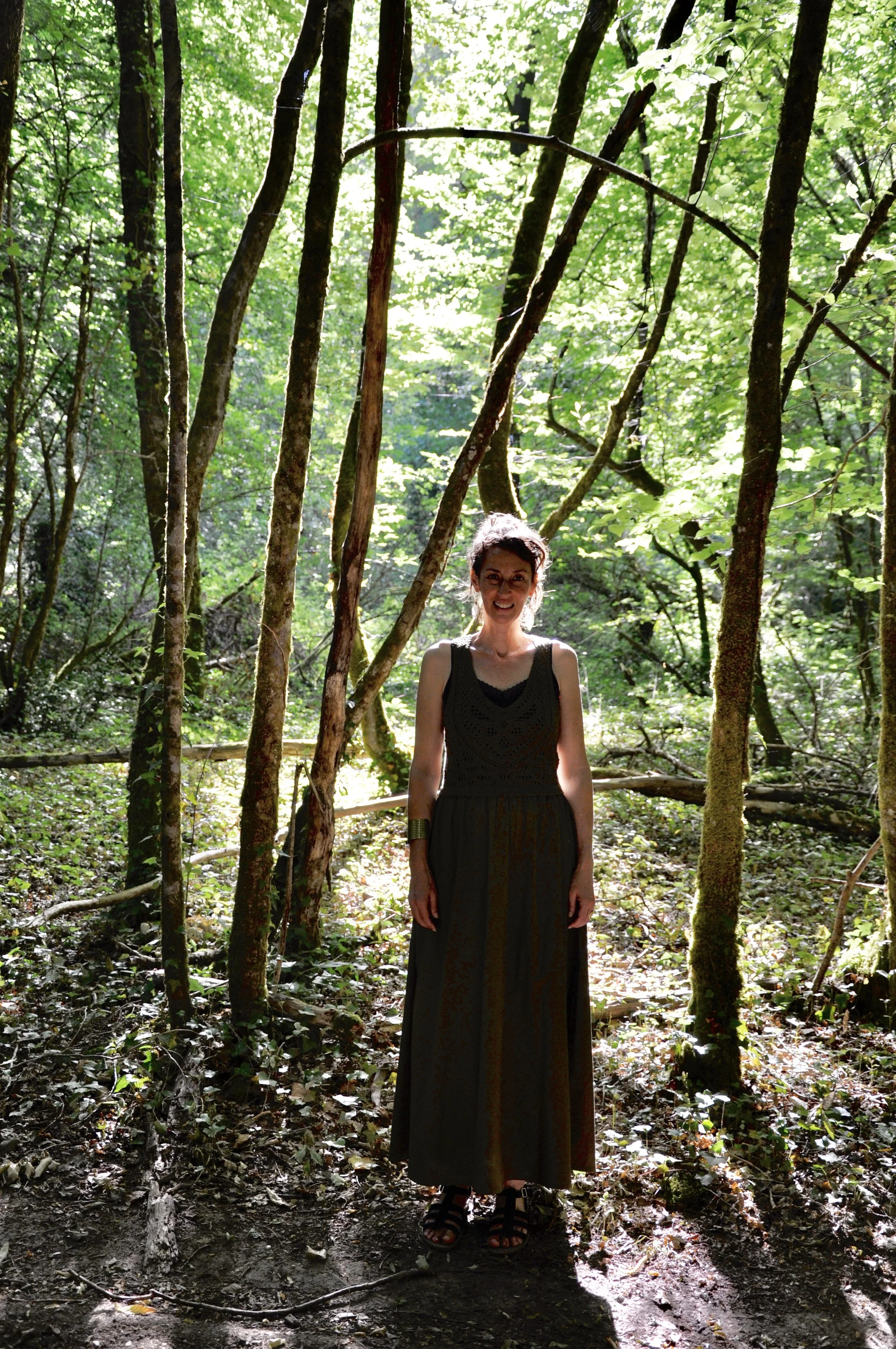 Une femme debout dans une forêt entourée d'arbres verts. Elle porte une robe longue noire et des sandales. La lumière du soleil filtre à travers les feuilles, créant des reflets lumineux dans la forêt dense.
