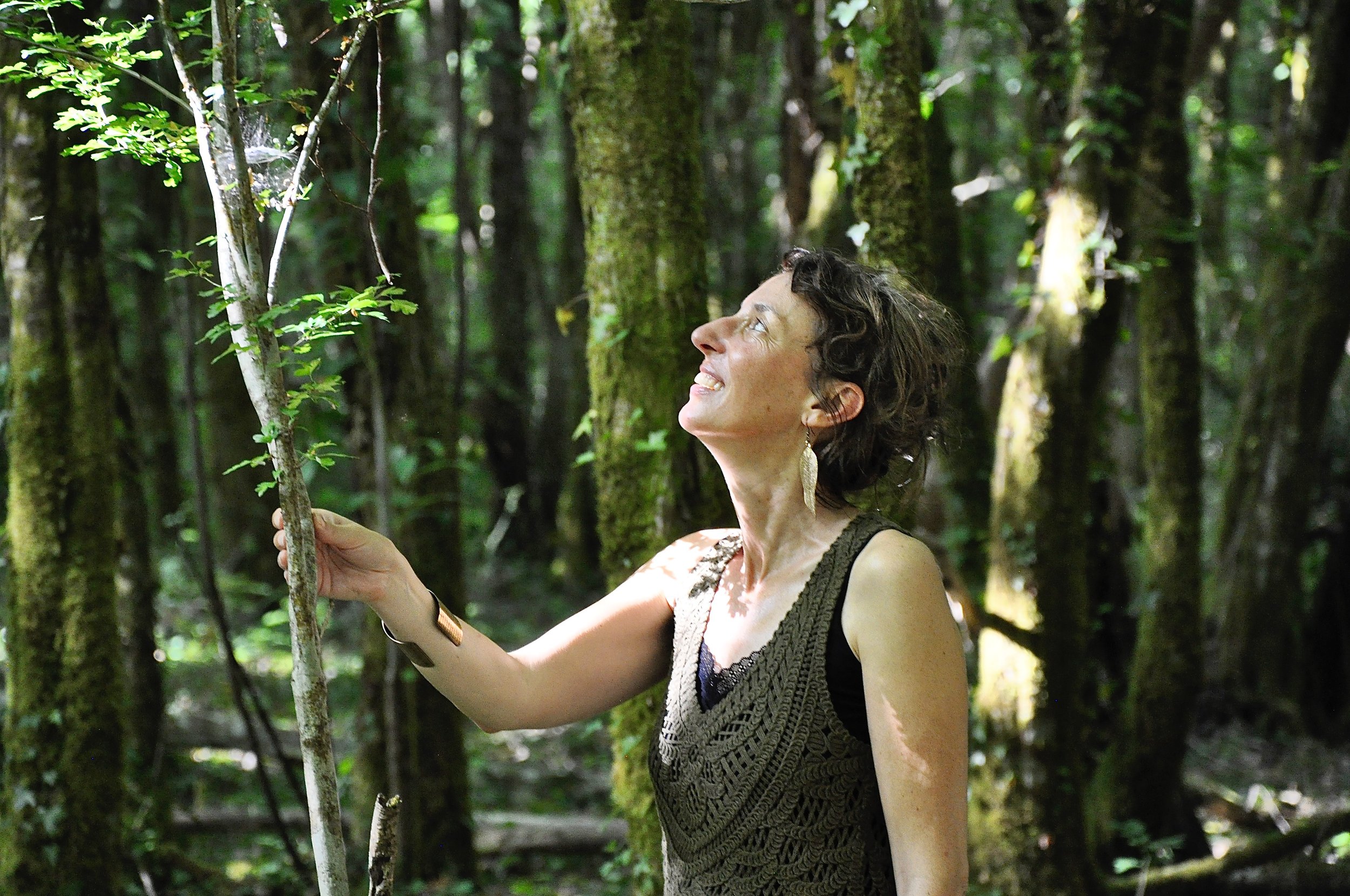 Une femme souriante dans une forêt dense, tenant un petit arbre ou une branche, entourée de végétation et d'arbres.