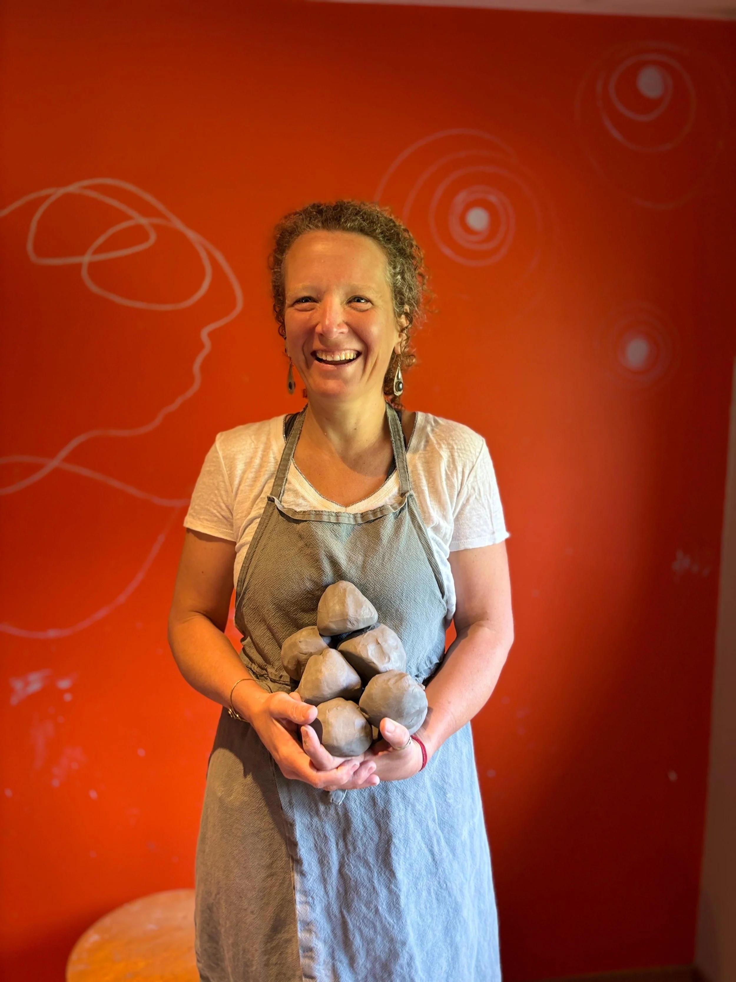 Femme souriante tenant une sculpture de plusieurs rochers en argile, portant un tablier gris clair, debout devant un mur orange avec des motifs circulaires blancs.