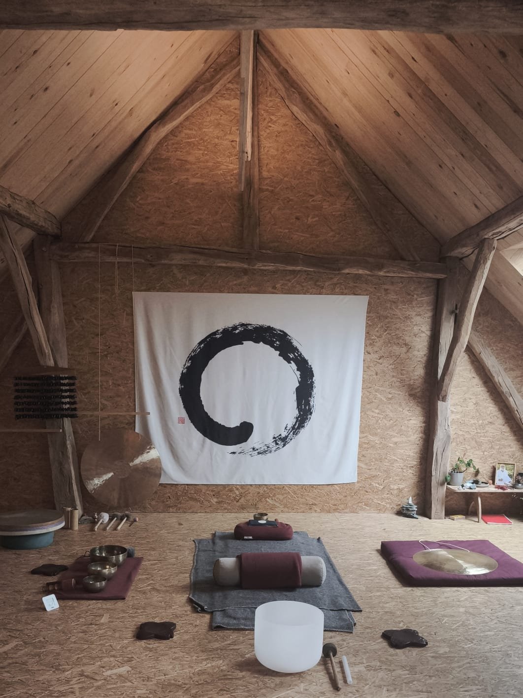 Salle de méditation zen avec décor minimaliste, tapis, bols tibétains, gong, et un large cercle noir sur un drapeau blanc suspendu au mur, dans un intérieur en bois chaleureux.