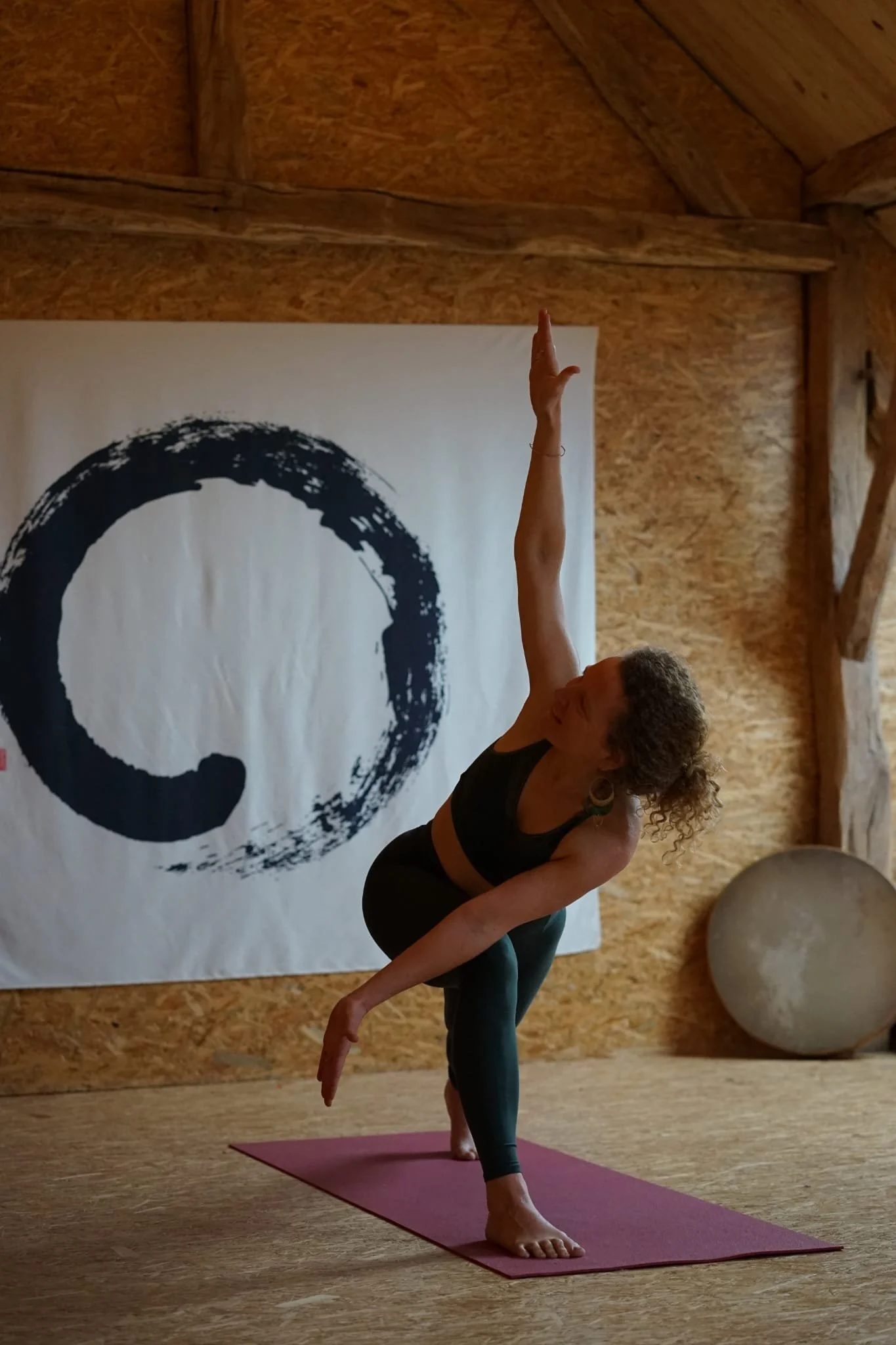 Une femme pratique le yoga sur un tapis rose dans une pièce en bois, en position d'équilibre avec un bras levé. Un tableau noir avec un cercle de calligraphie japonaise est accroché au mur.