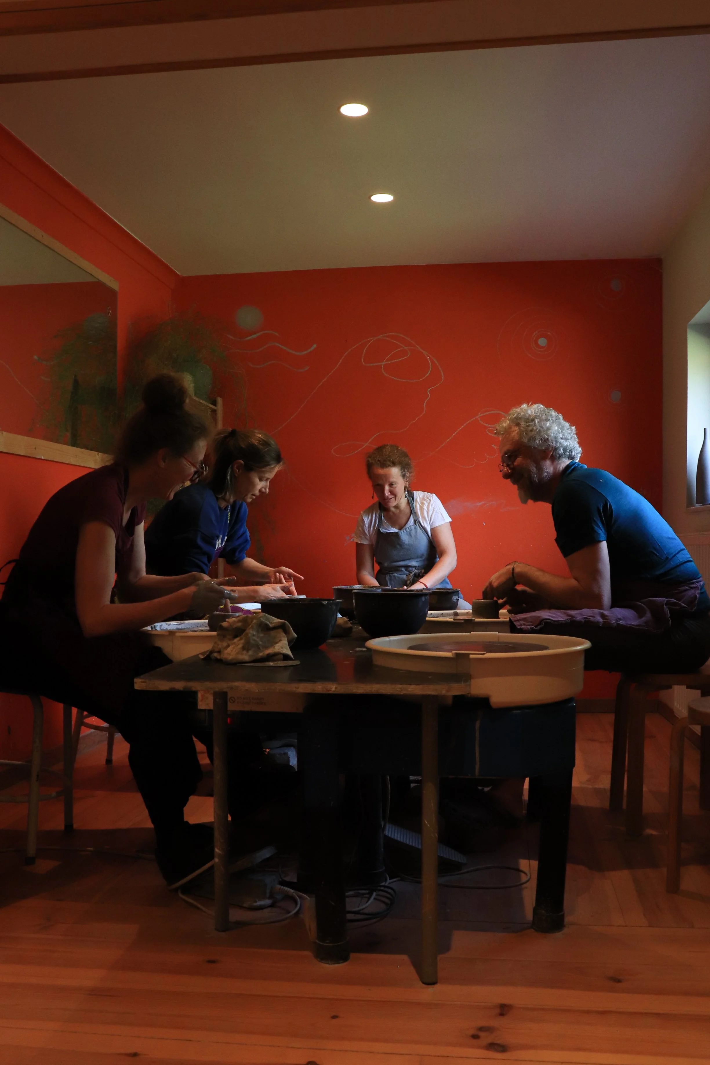 Cinq personnes participent à un atelier de poterie dans une pièce avec un mur orange décoré de dessins de formes géométriques et de lignes blanches. La scène est photographiée en intérieur, avec un plafond éclairé par des spots lumineux.