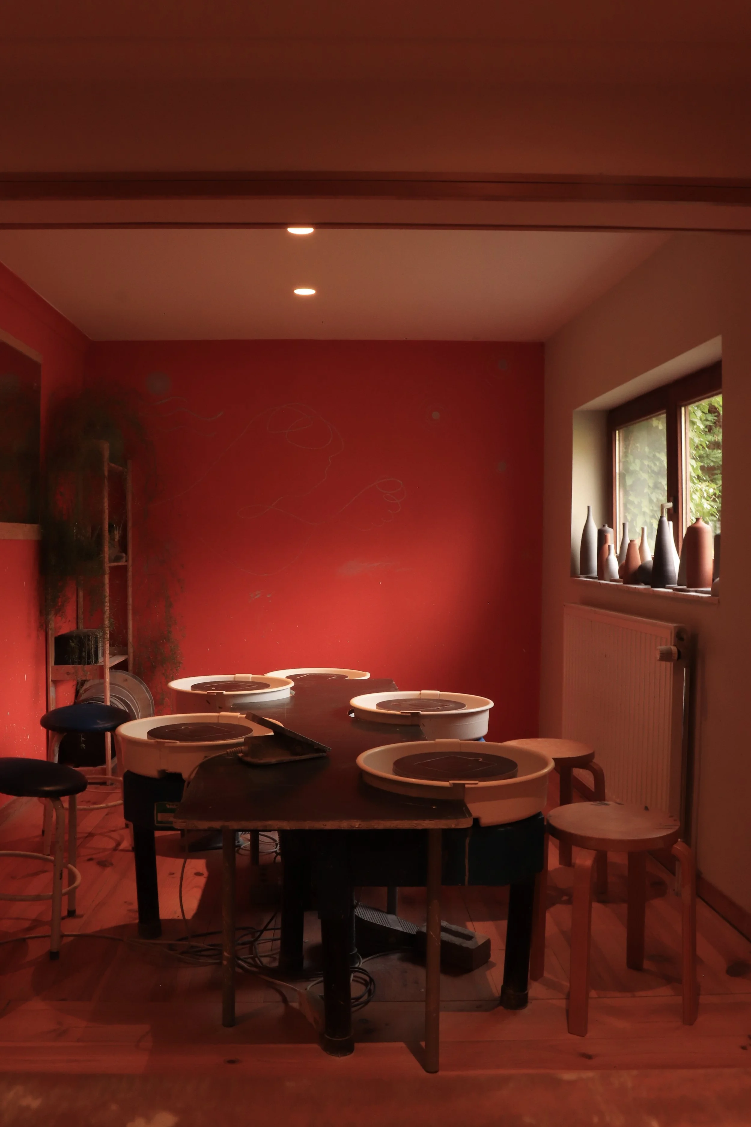 Une pièce avec un mur rouge, une fenêtre avec des plantes vertes à l'extérieur, une table ronde avec des tapis de cuisson pour enfants, et plusieurs chaises en bois.