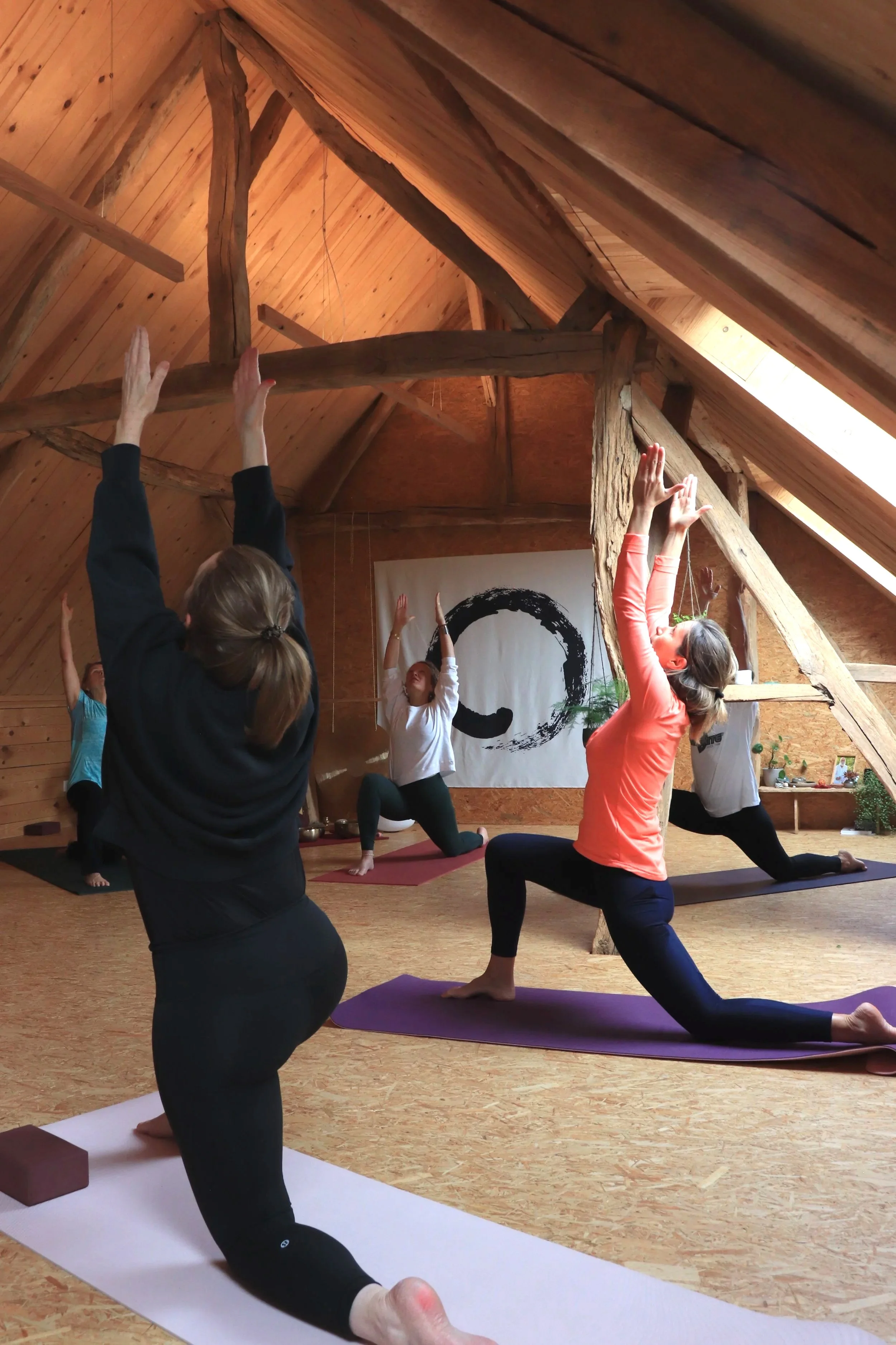 Des personnes pratiquent le yoga dans un salon en bois, en position de guerrier, avec un grand symbole noir d'encre en toile de fond.