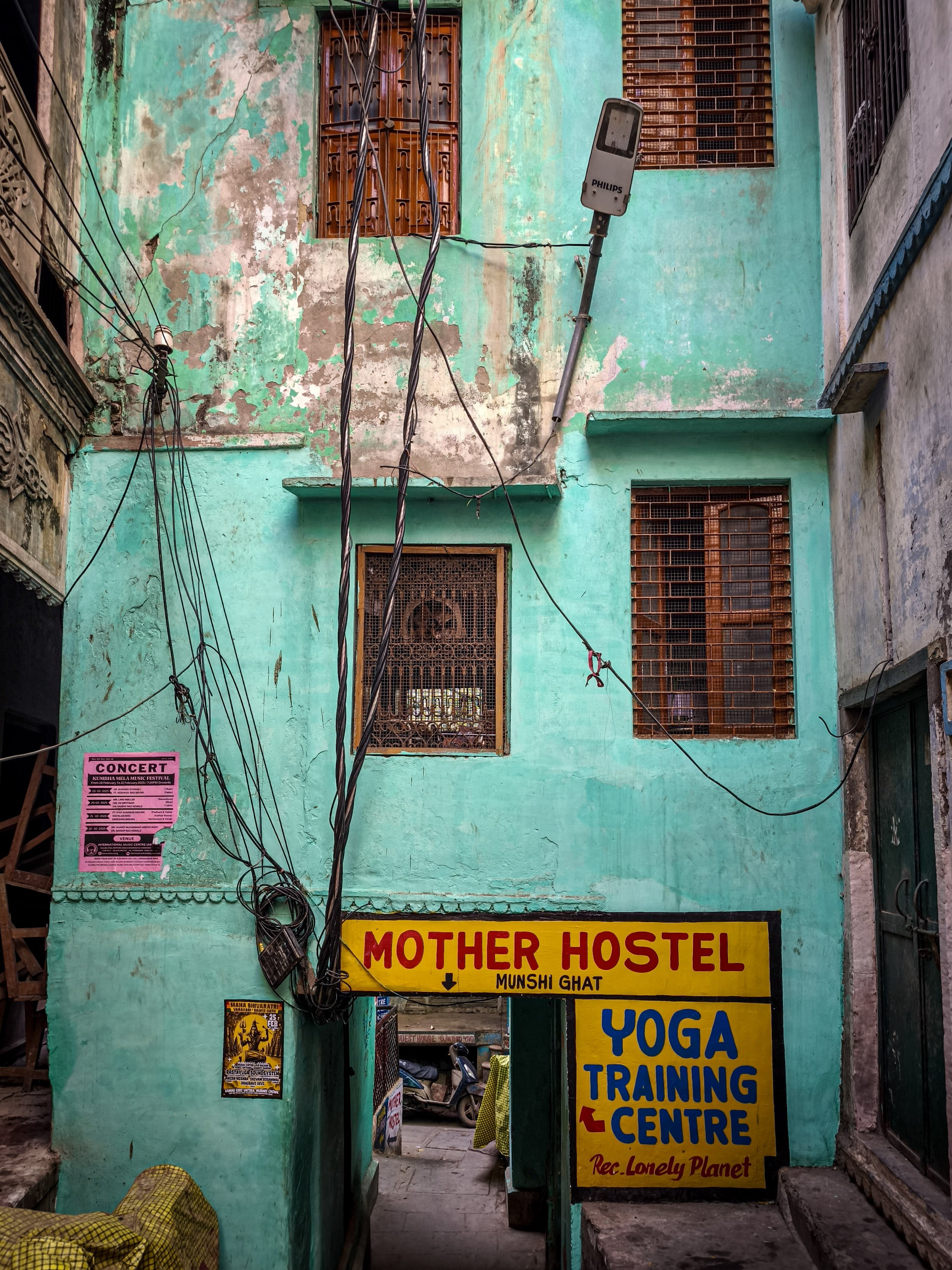 Vue d'une entrée de chambre d'auberge ou centre d'entraînement avec des fenêtres en métal et un mur vert turquoise délavé. Il y a plusieurs fils électriques suspendus, une affiche rose pour un concert, et une grande fiche signalétique jaune avec du texte en anglais indiquant un centre d'entraînement de yoga.