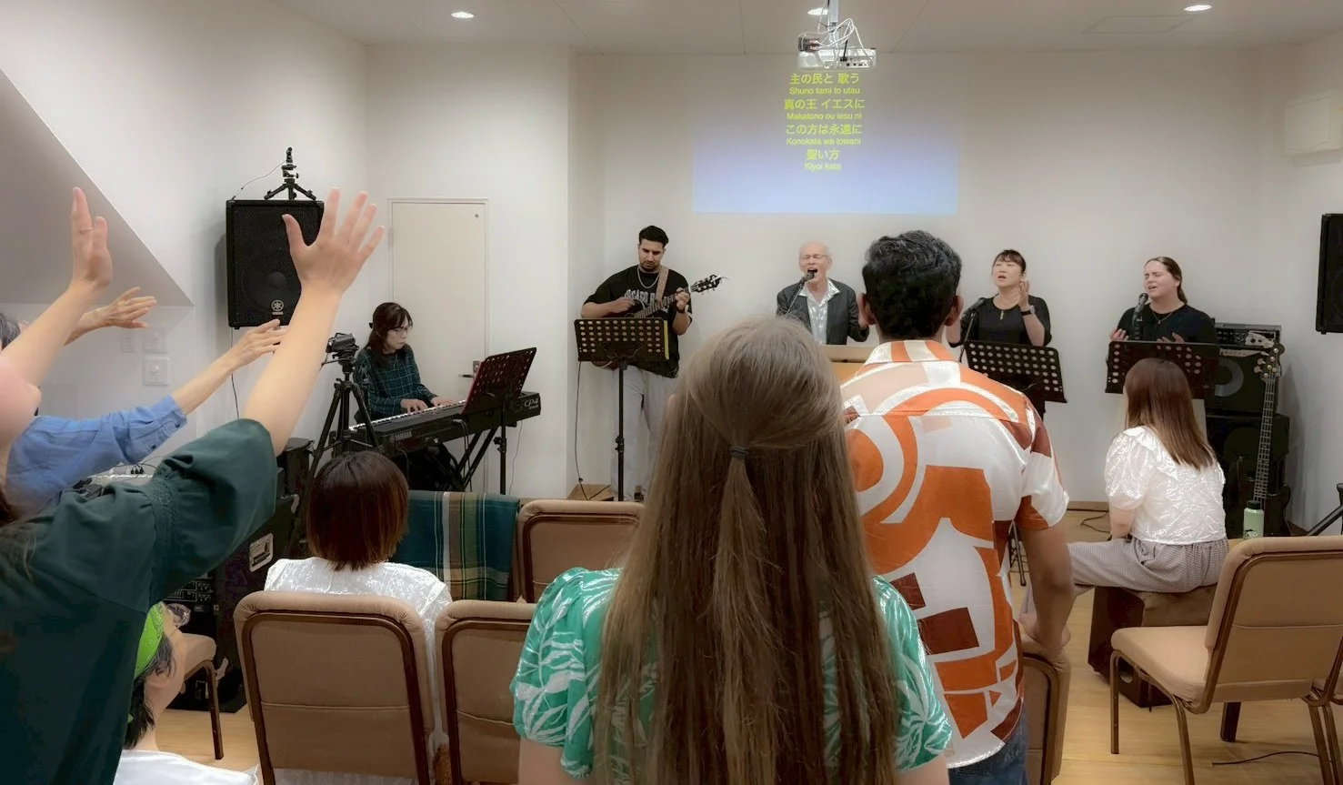 Group of people singing with musicians in a small room, some audience members raising hands, a woman playing keyboard, a man playing guitar, and a man singing with others accompanying in a church or community setting.