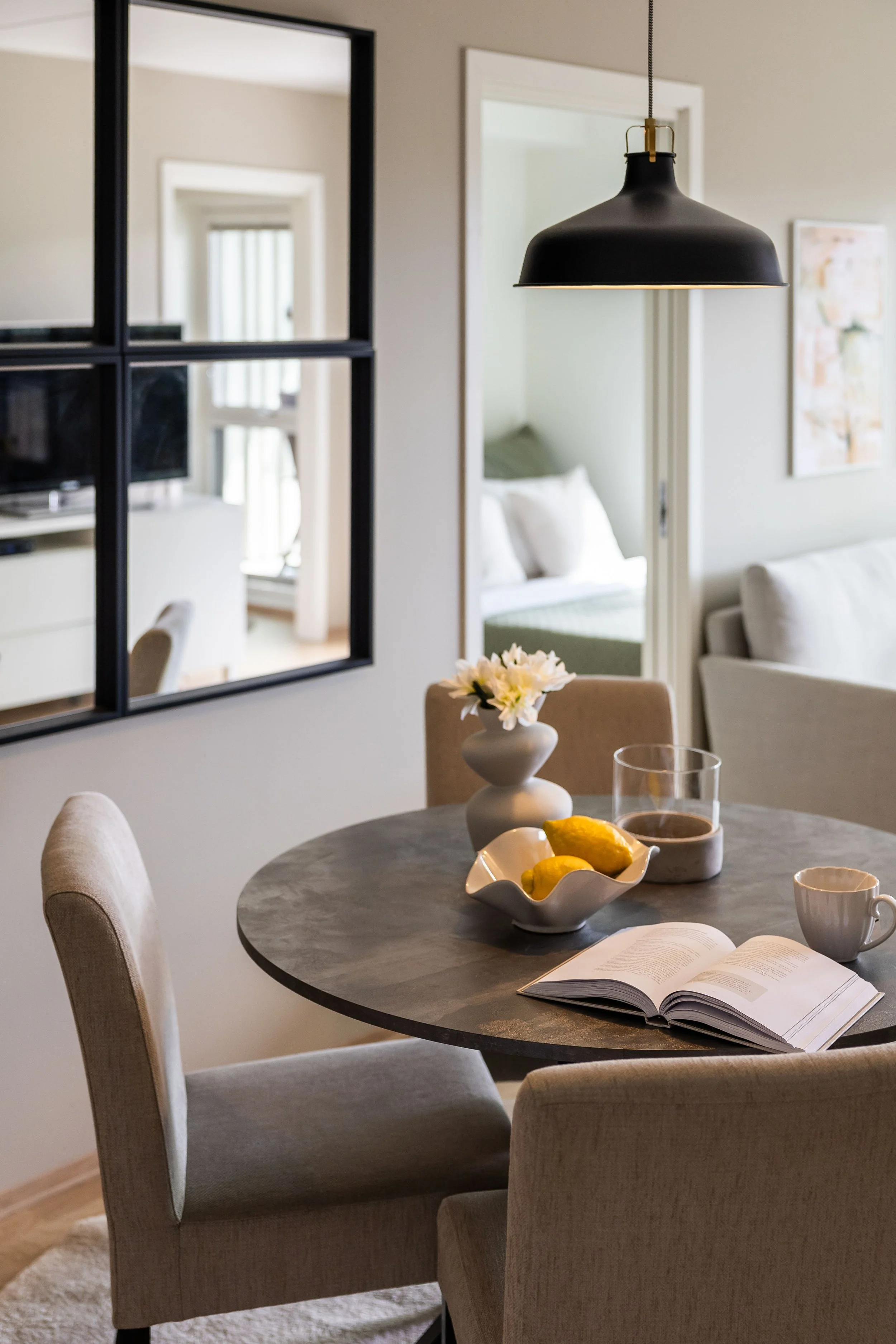Et rundt spisebord med beige stoler, en åpen bok, en kopp, en skål med sitroner, en vase med hvite blomster, og et dokument. På veggen henger et svart rammet speil, og i bakgrunnen kan en stue med sofa og et soverom være synlig.