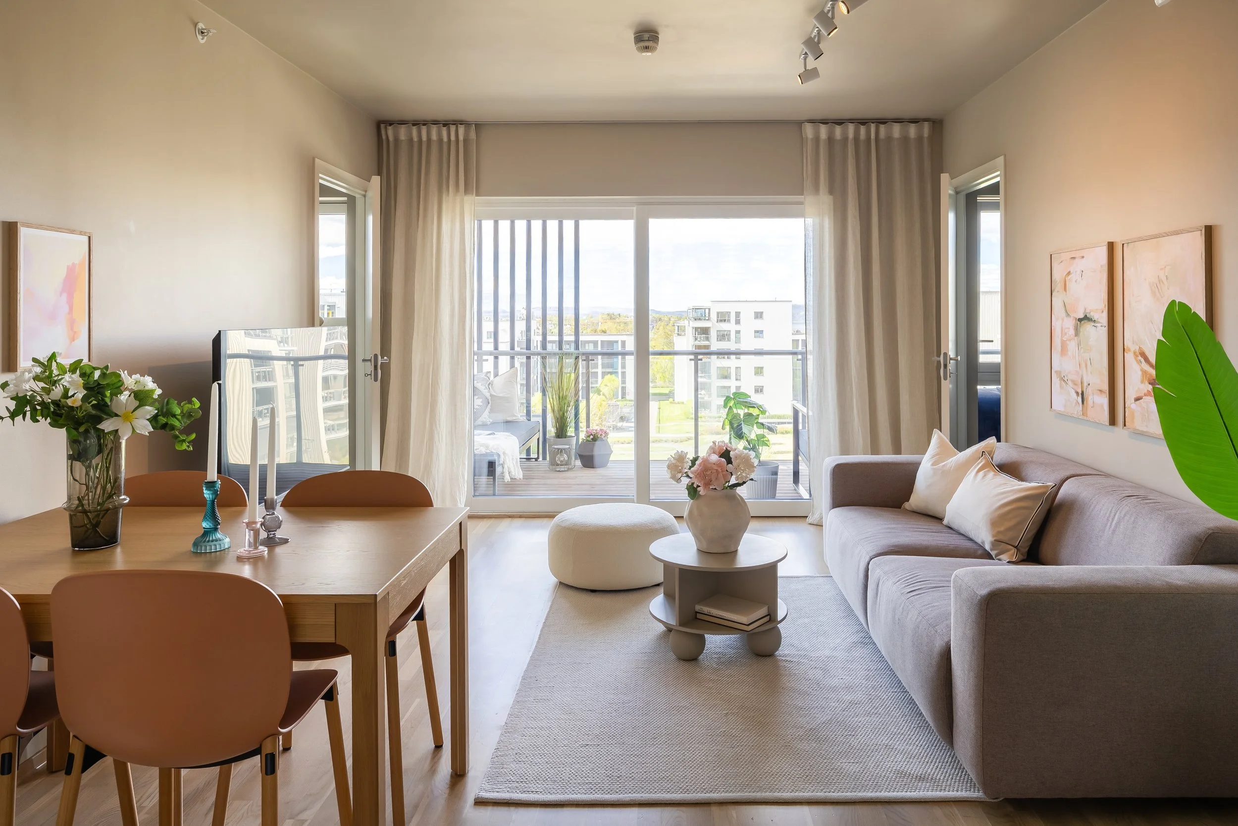 Et lyst og innbydende stueområde med en stor sofa, et lite rundt bord med blomster, og en stor glassdør som fører ut til en balkong med landsbyutsikt. Innredningen inkluderer veggdekorasjoner, grønne planter og gardiner.