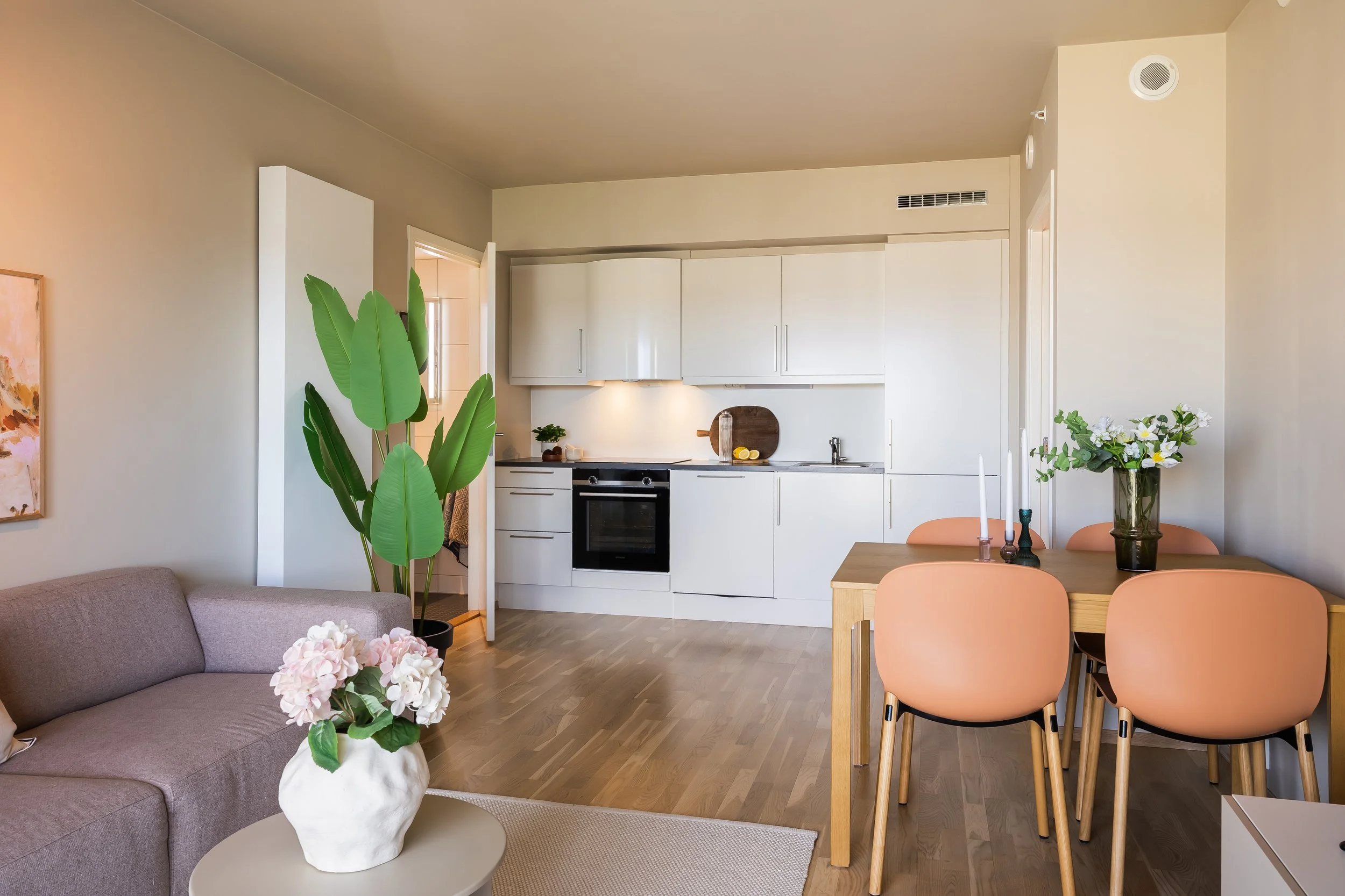 Et moderne, lyst kjøkken med hvite skap og integrert ovn, ved siden av en spisestue med fire rosa stoler rundt et trebord. Det er planter og blomsterdekorasjoner i rommet, inkludert en stor grønn plante og en vase med hvite blomster.