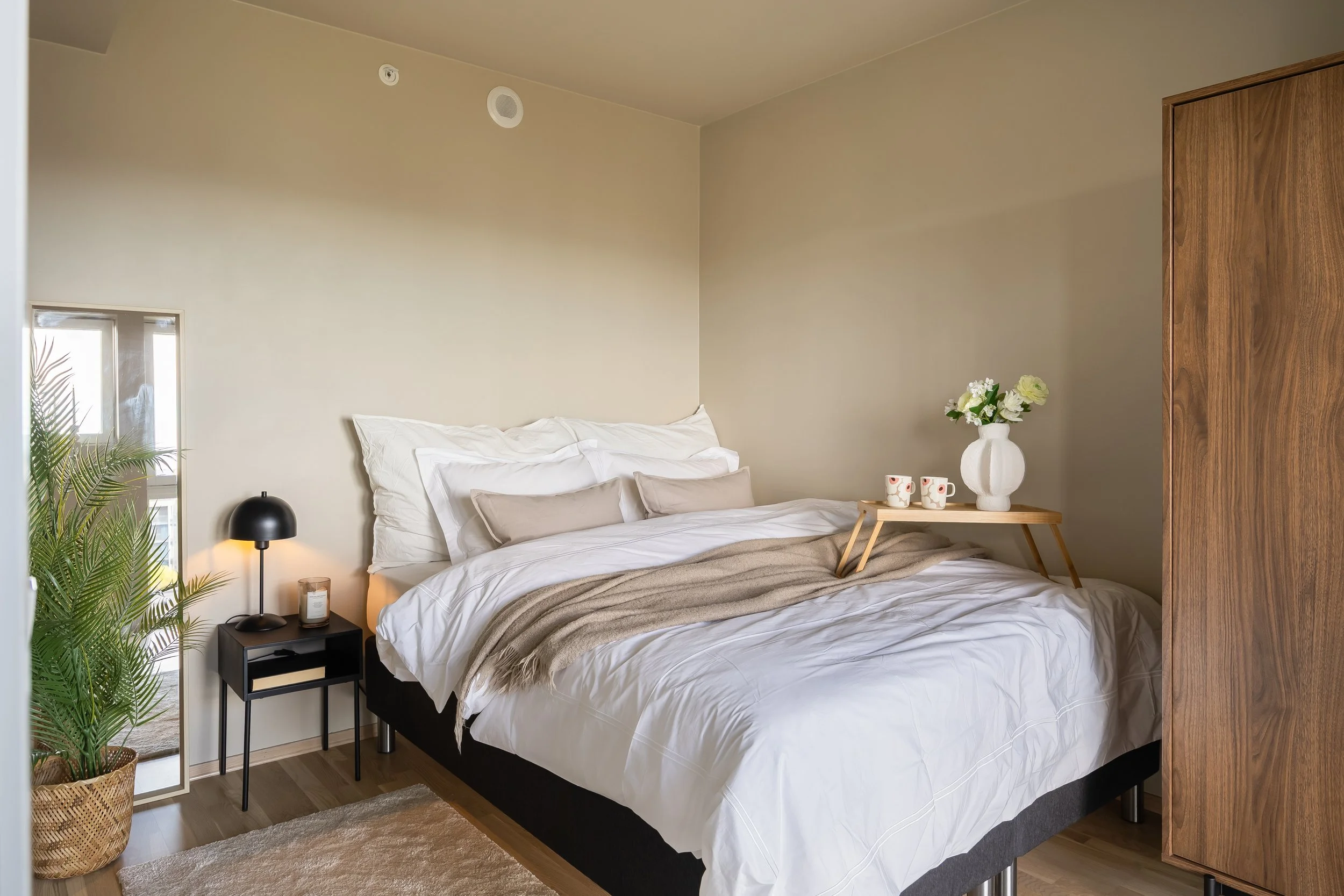 Soverom med seng, nattbord, lampe, blomster, speil og garderobe.