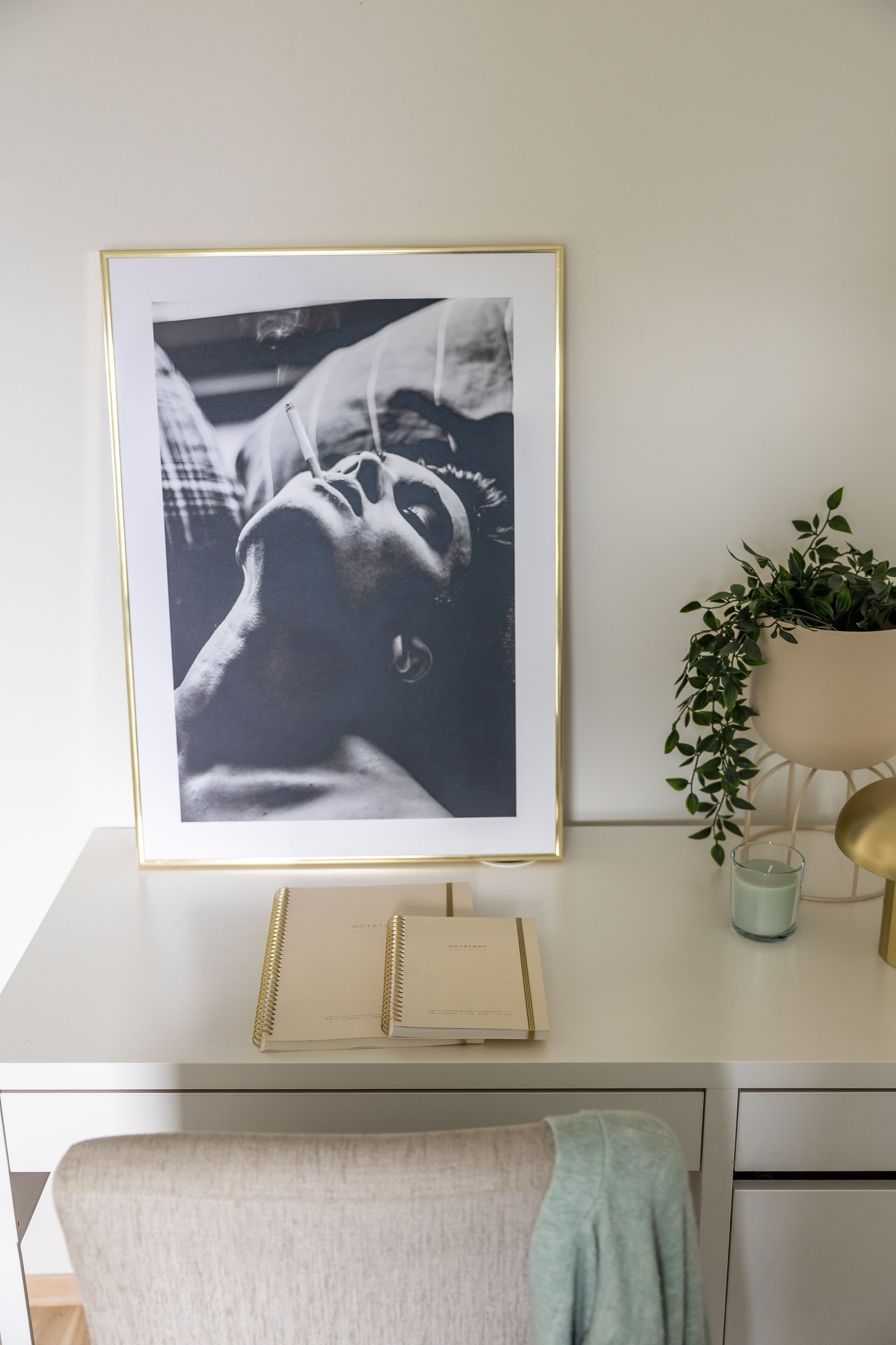 Et skrivebord med en stor, vertikal svart-hvitt bilde av en person som ligger ned med en sigarett i munnen, omgitt av planter, notatbøker og en liten lysestake.