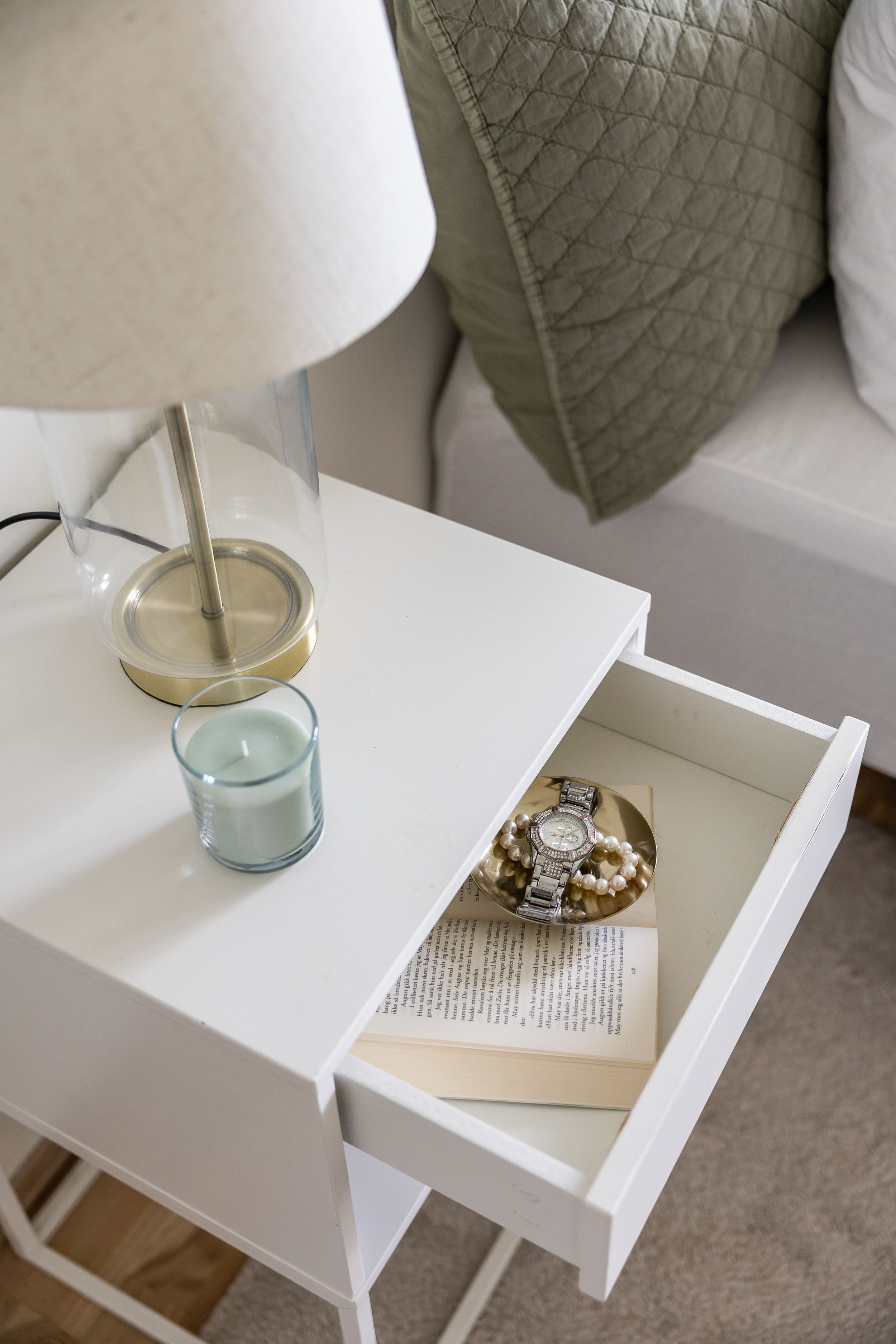 Nært bilde av en hvit nattbord med en glasslampe, to lyslykter, et armbåndsur og perlearmbånd på en skål, samt en åpen bok under nattbordet. Loftlampe i forgrunnen og seng med mørkegrå sengesjal og puter i bakgrunnen.