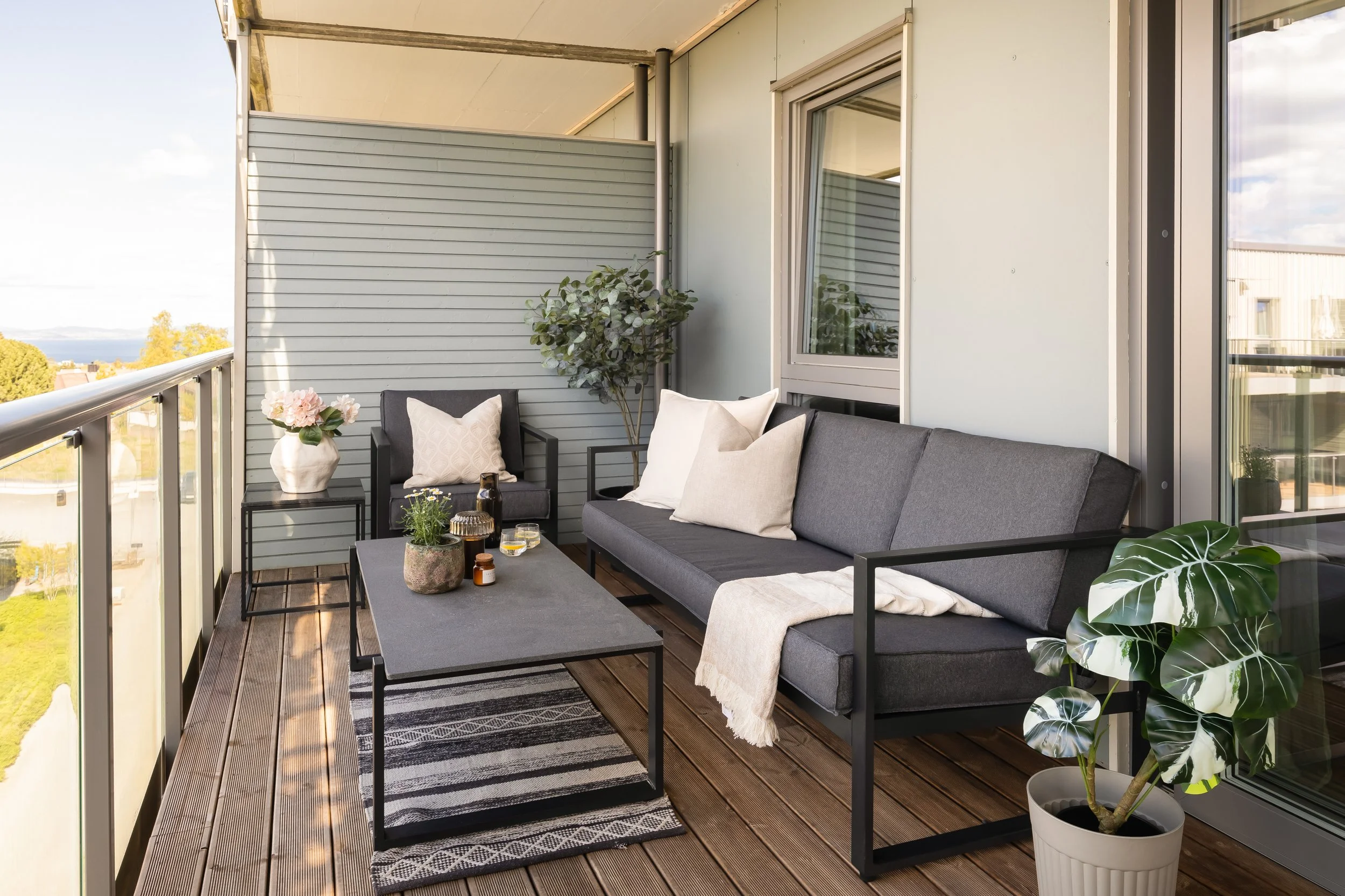 Terrasse med utendørs møbler, inkludert sofa, stol og bord med blomsterpotte, omgitt av glassrekkverk, med utsikt over trær og bygninger i bakgrunnen.