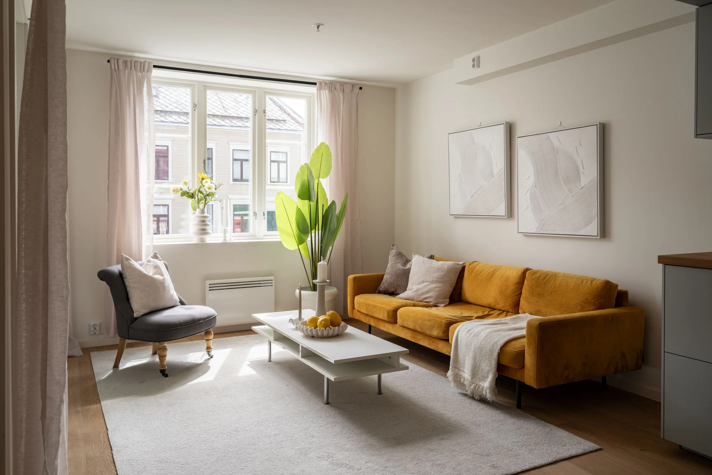 Stue med stor sofa i sennepsgul, to puter, et runde lagerskap med frukt, stor grønn plante, vindu med lyse gardiner, to veggkunstverk.